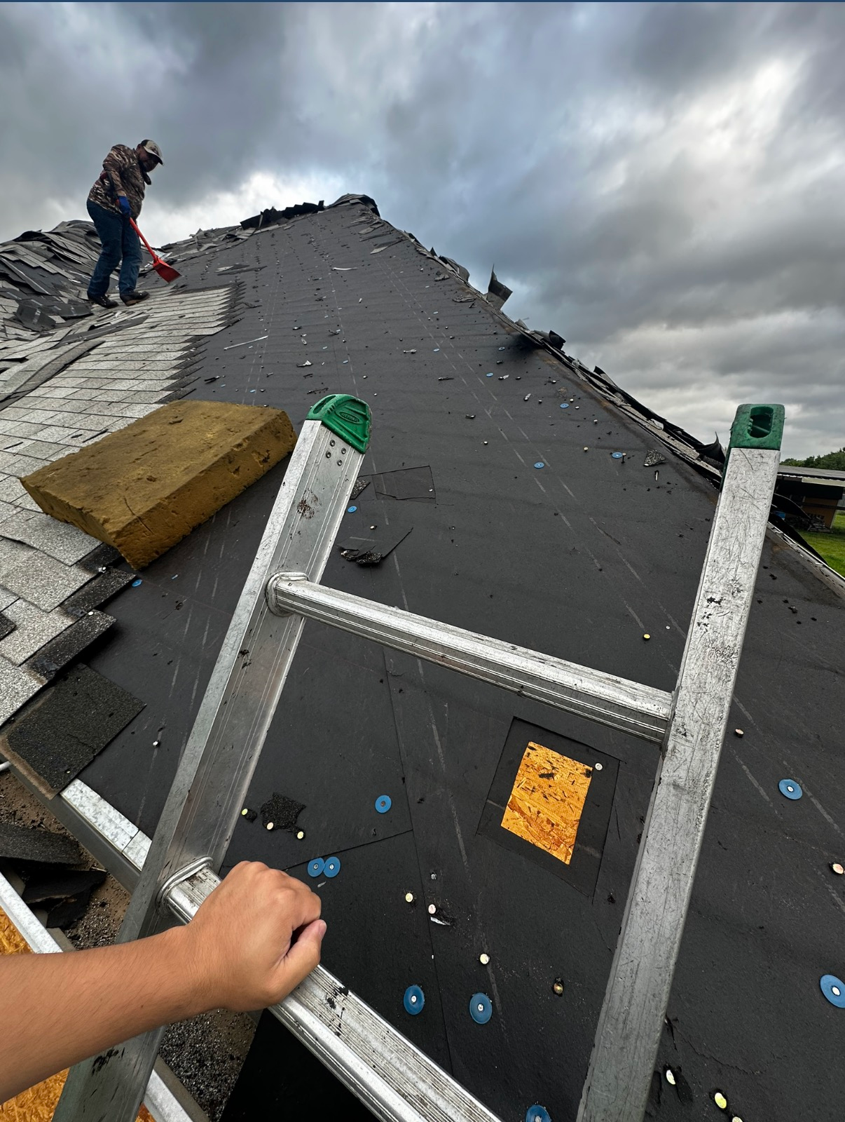 A person standing on a ladder holds onto it while viewing a worker removing shingles from a residential roof.