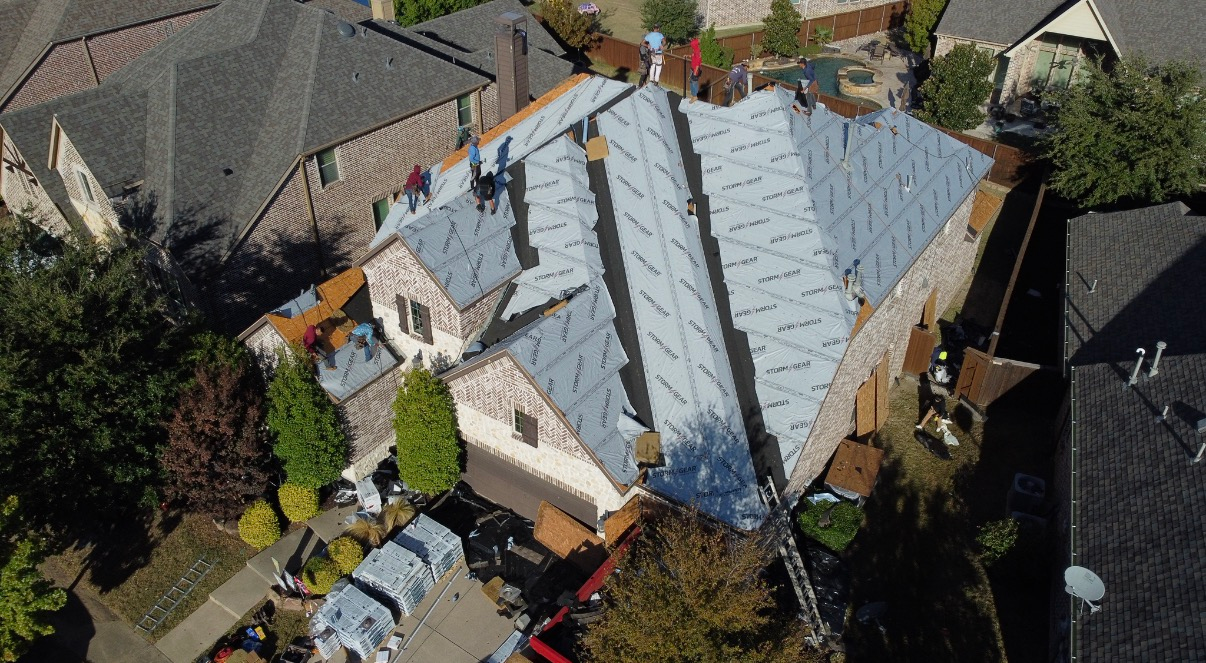 An aerial view of workers installing light-colored underlayment on a residential roof in a suburban neighborhood.