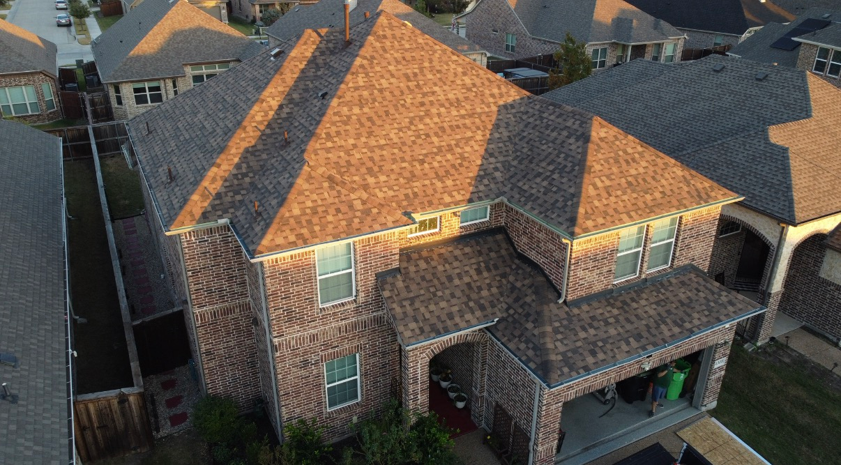 Aerial view of a two-story brick house with a brown shingled roof, surrounded by neighboring homes in a suburban area.