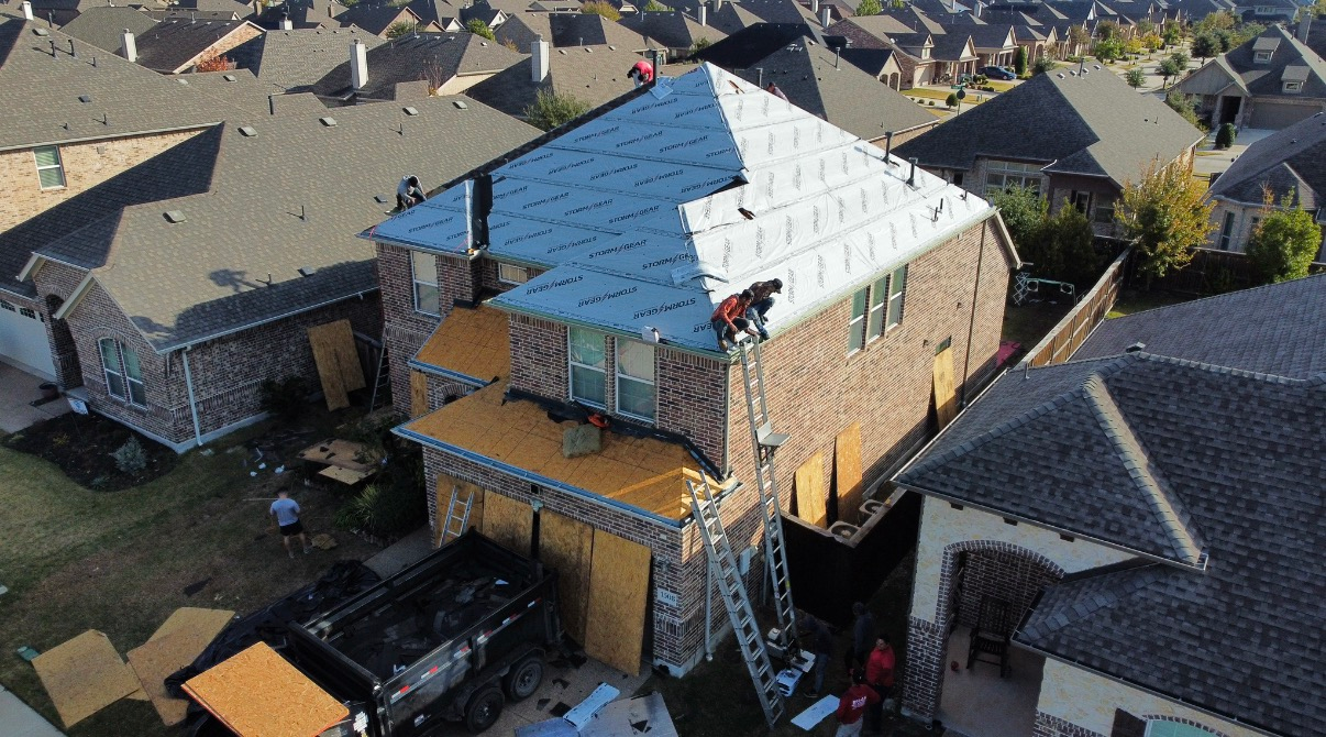 Workers on a roof, installing underlayment on a residential home with a dumpster in the driveway below.