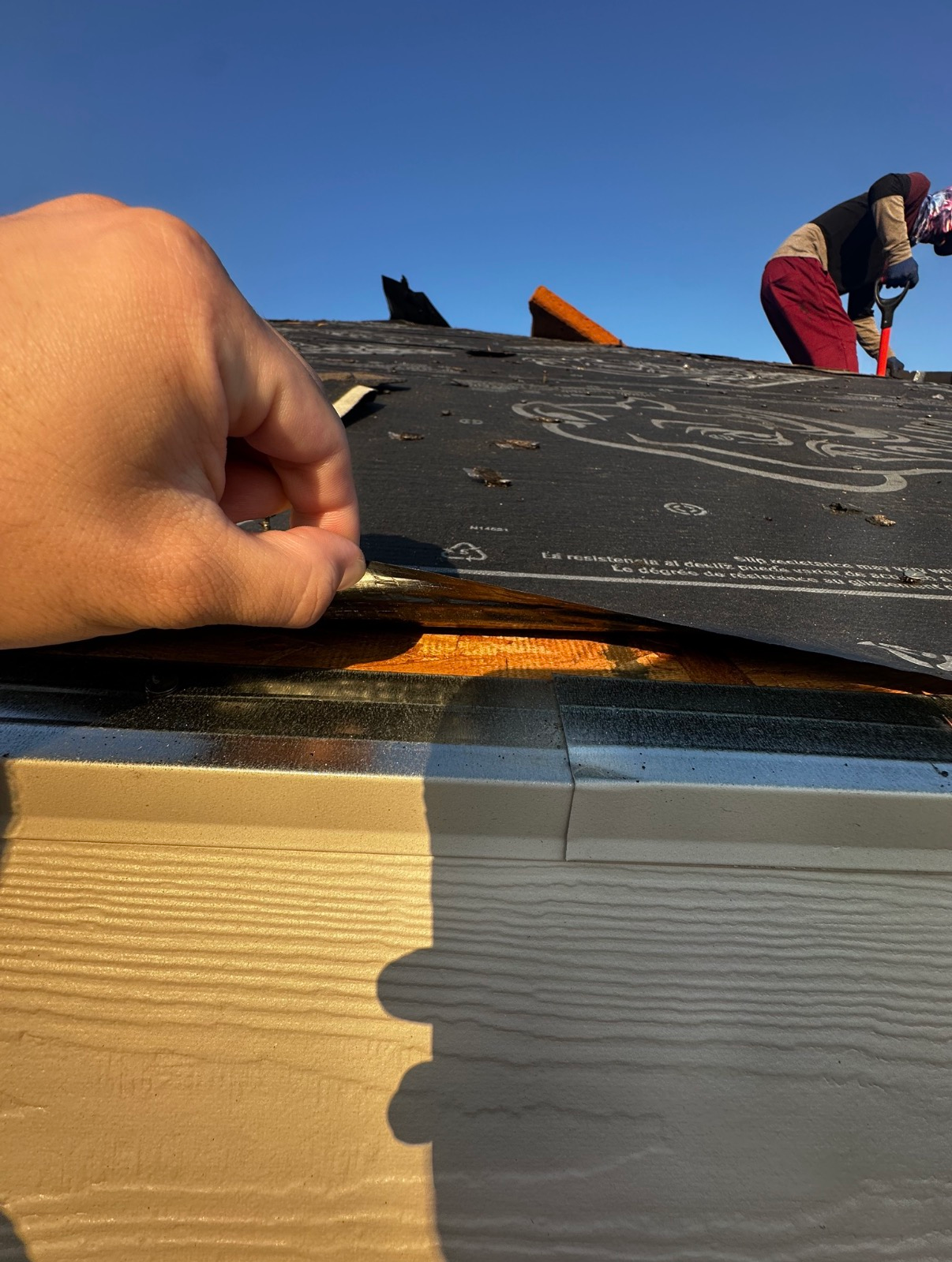 A person lifting the edge of black roof underlayment to reveal the wood decking, with a roofer working in the background.