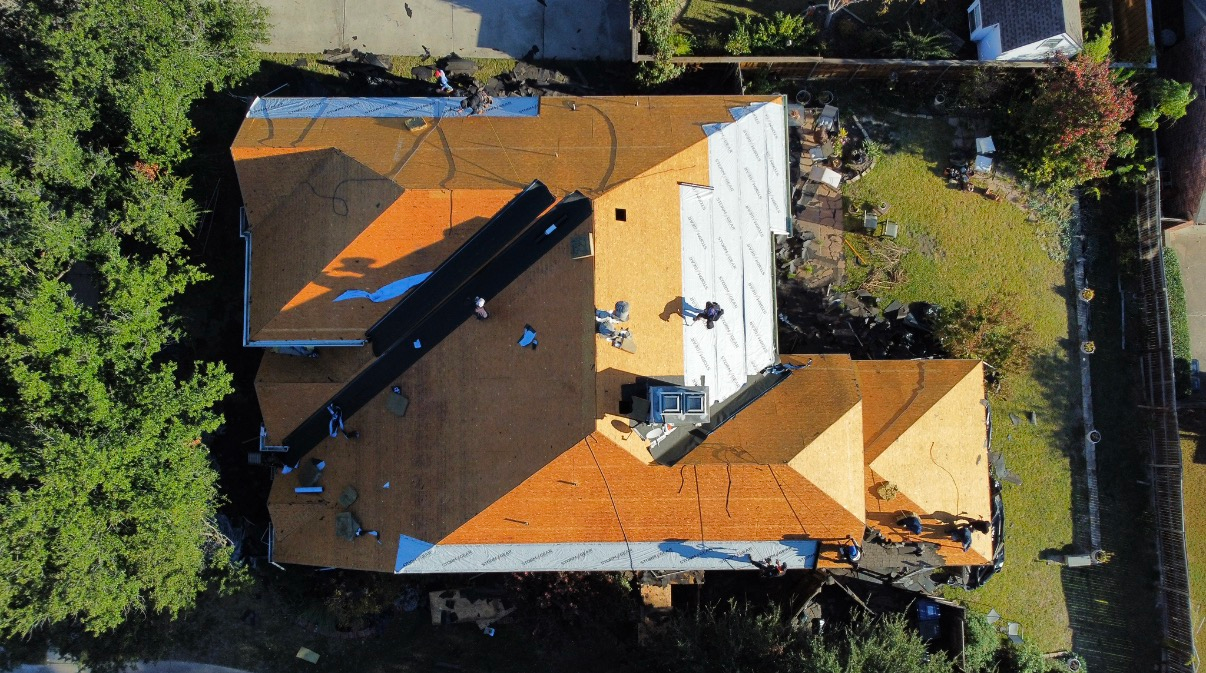 An aerial view shows a residential roof undergoing repair with sections of plywood sheathing and white underlayment.