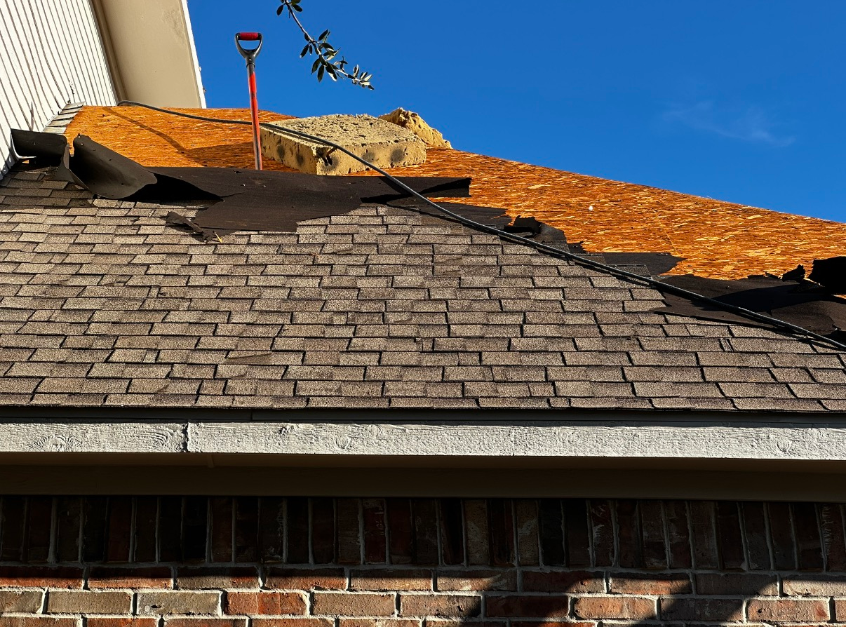 A roof under repair, showing torn shingles exposing the plywood deck with a shovel standing against the upper section.