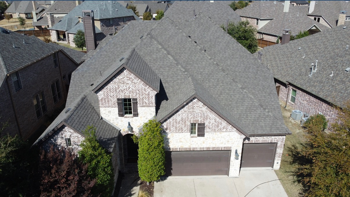 Aerial view of a two-story residential brick house with a gray shingled roof, a multi-car garage, and driveway.