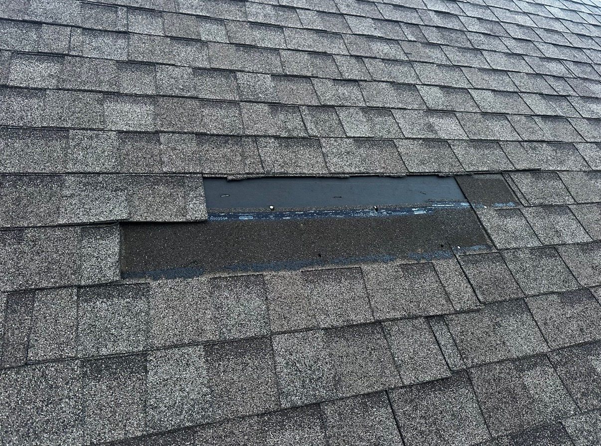 A missing rectangular patch of gray asphalt shingles on a roof, revealing the black underlayment beneath.
