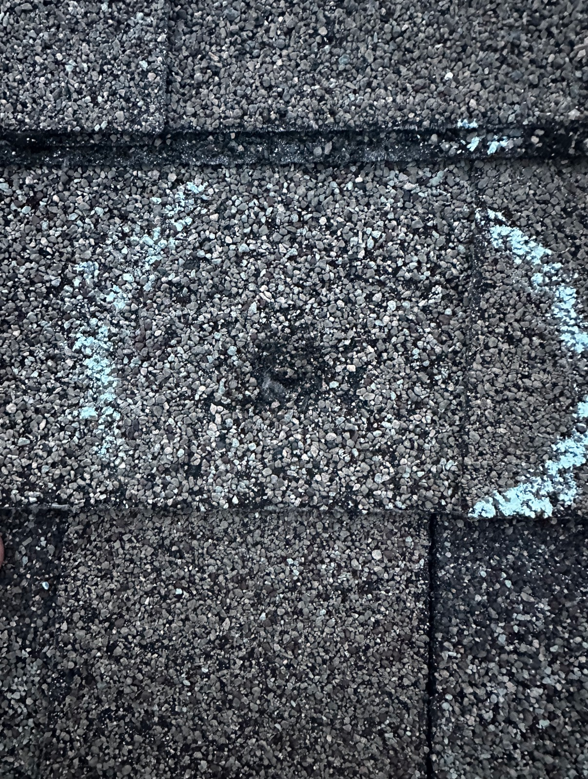 A circular white chalk mark drawn on a textured gray asphalt roof shingle, highlighting a small puncture or impact point.
