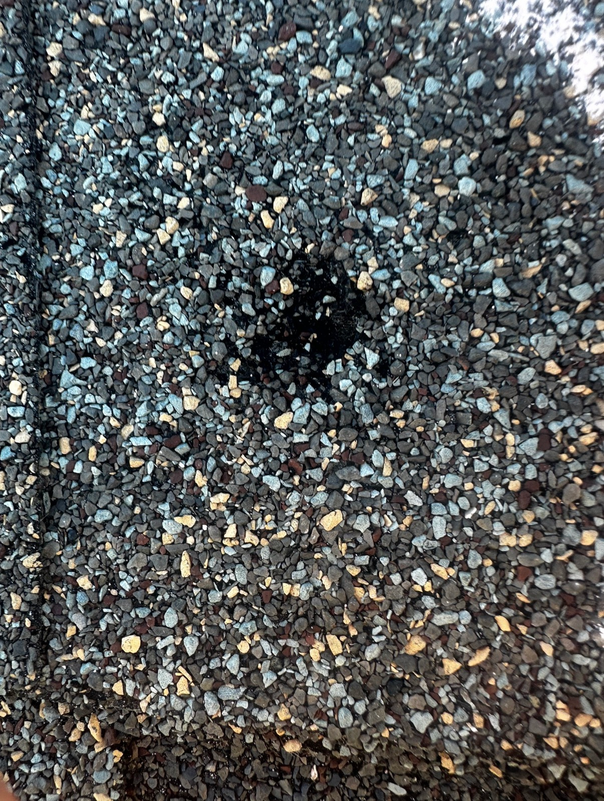A close-up view of asphalt roofing shingles with a small patch of missing surface granules, exposing the black base layer.