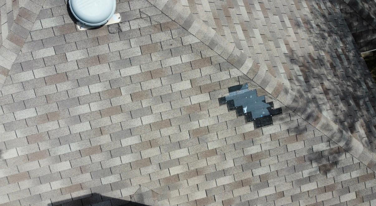 An aerial view of a tan shingled roof with a white circular ventilation cap and a grey rectangular roof vent.