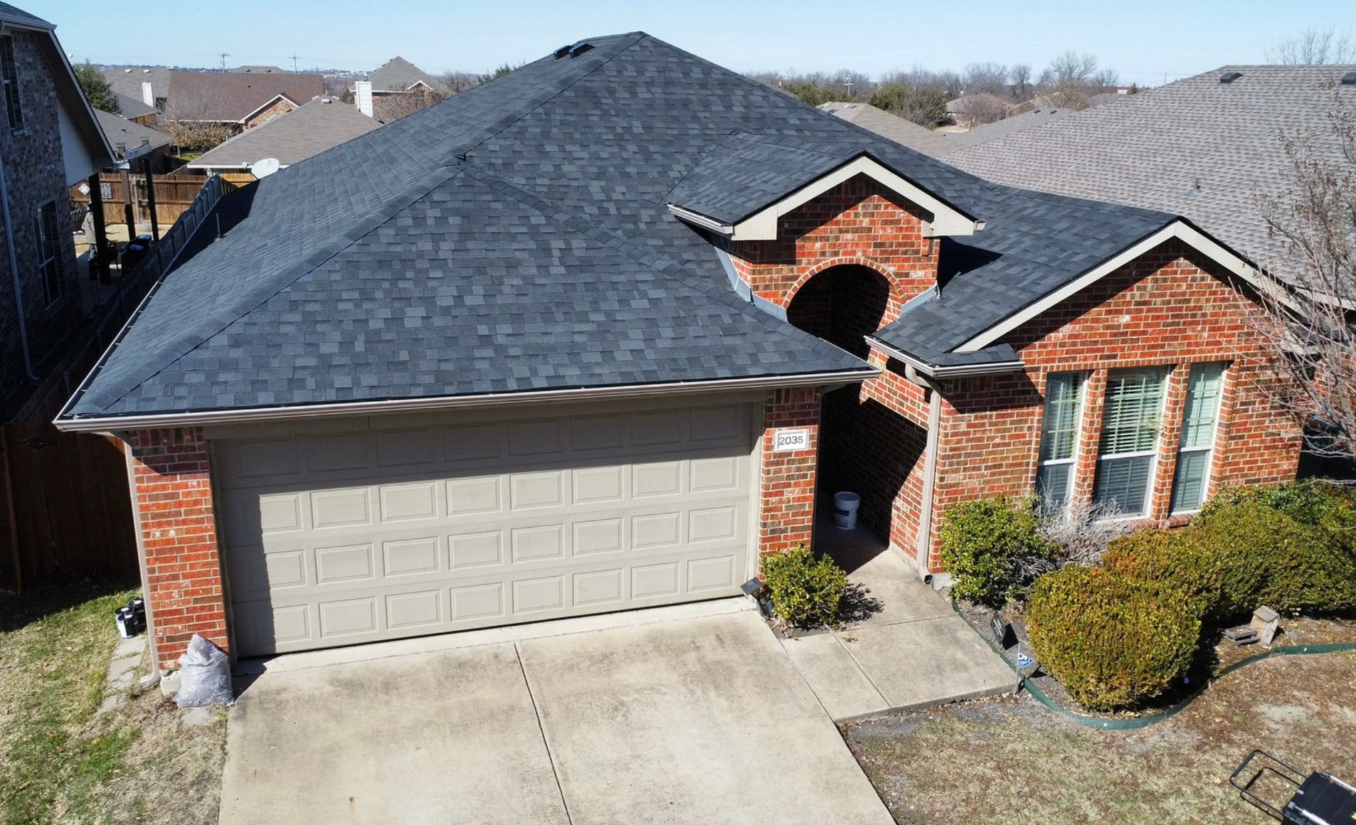 A high-angle view of a brick house with a dark shingled roof, a two-car garage, and a small front yard with shrubs.
