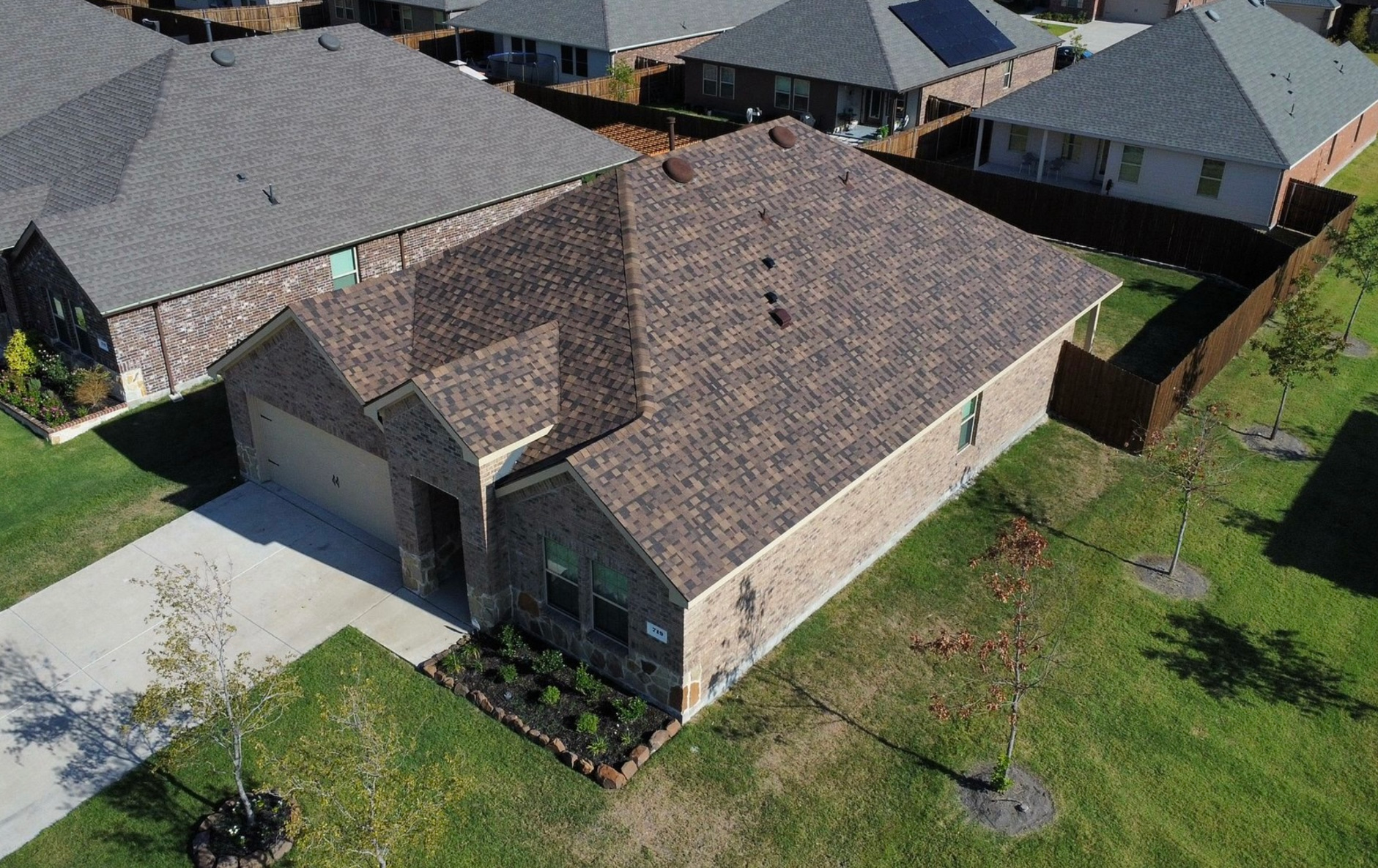 High-angle view of a brick house with a dark shingled roof, a concrete driveway, and a lawn in a suburban neighborhood.