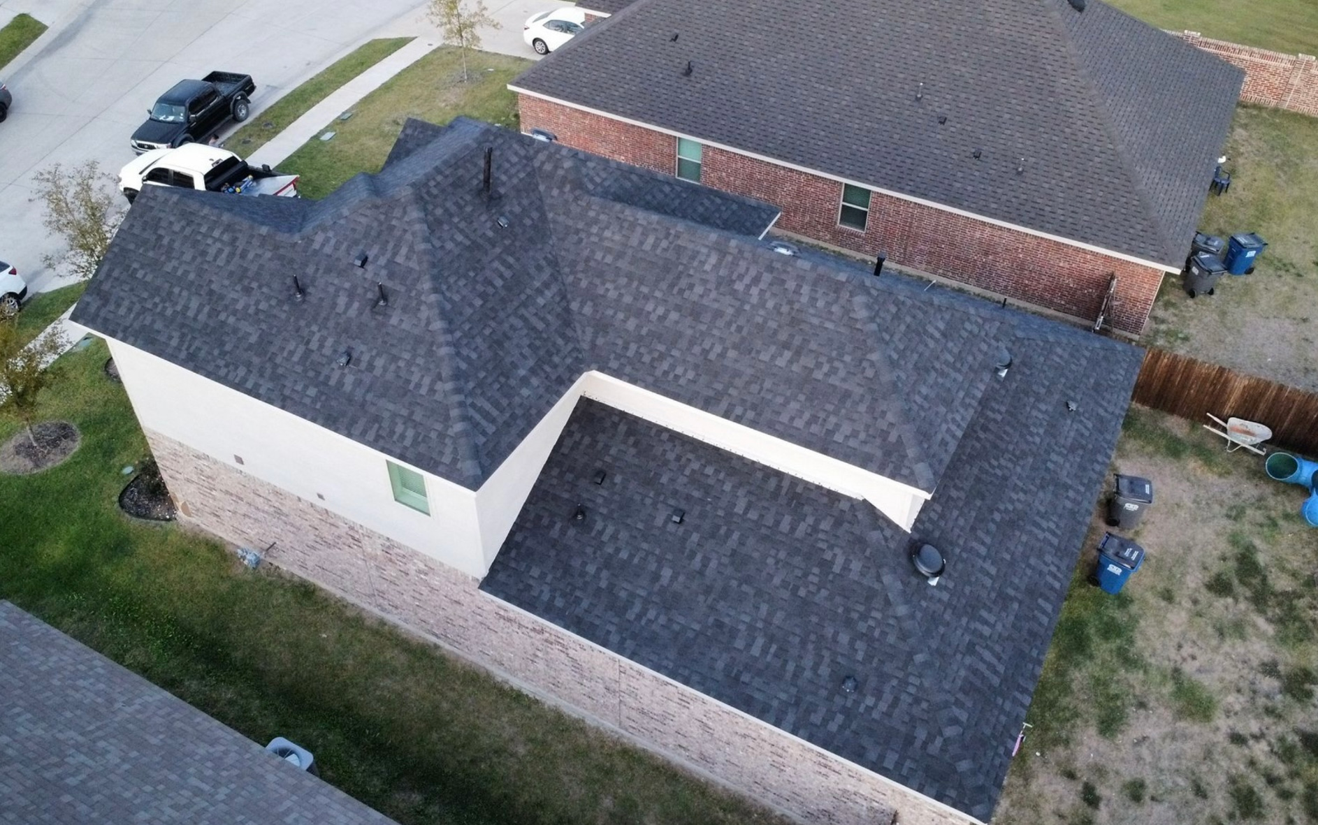 An aerial, high-angle view of a residential house with a gray shingled roof, next to a brick-faced home.