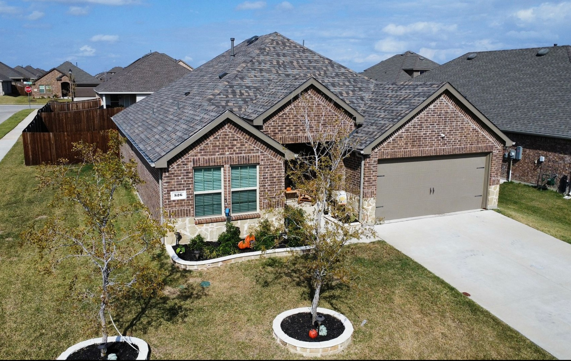 A suburban, single-story brick house with a gray shingled roof, two-car garage, and a manicured front lawn under a blue sky.