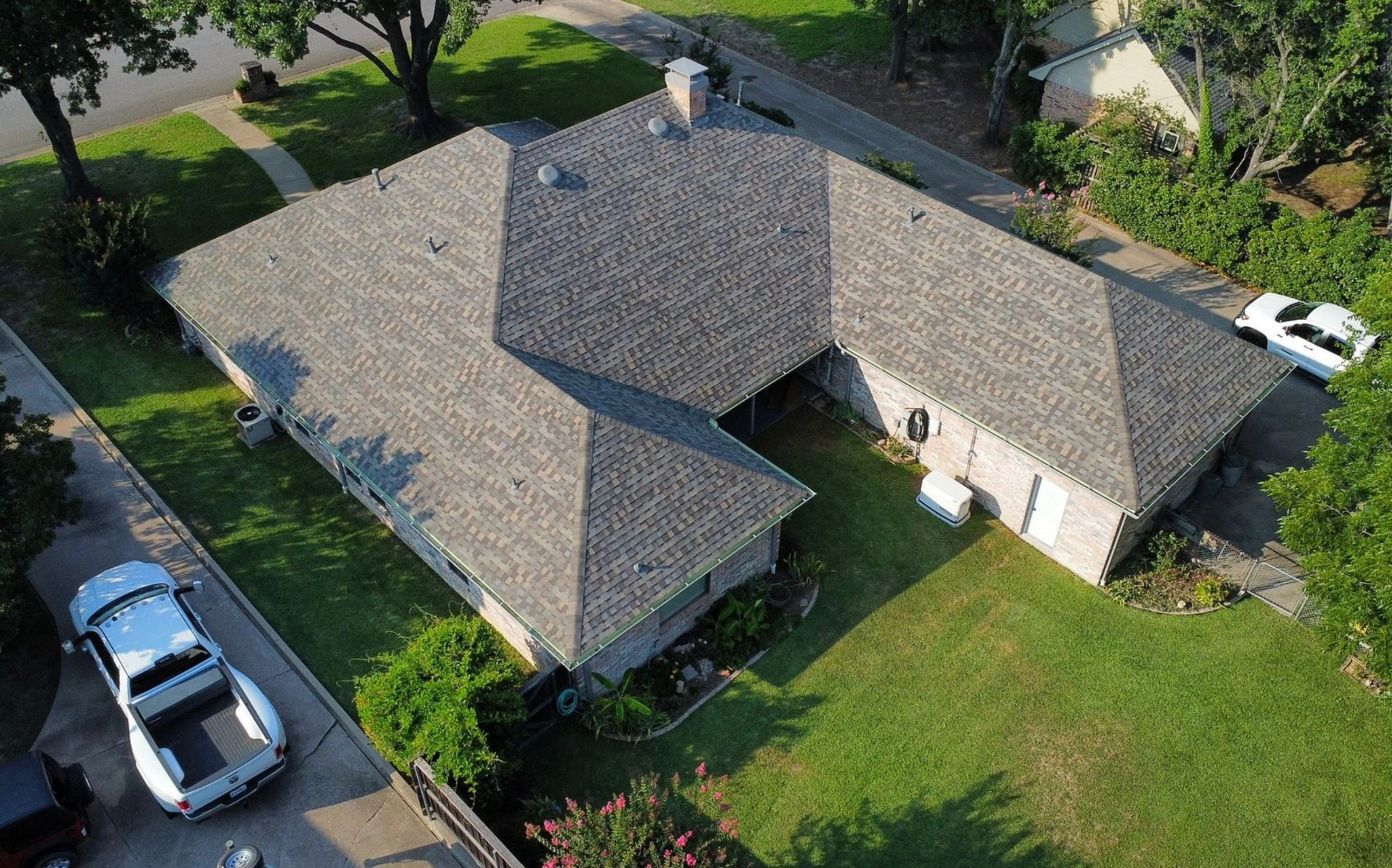 An aerial view of a single-story house with a grey shingled roof, surrounded by a lawn and parked vehicles.
