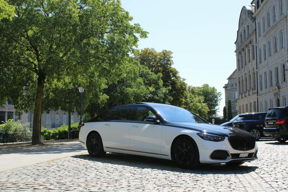 Une berline de luxe noire circule dans une rue ensoleillée, près de murs de pierre et d'arbres, en milieu urbain.