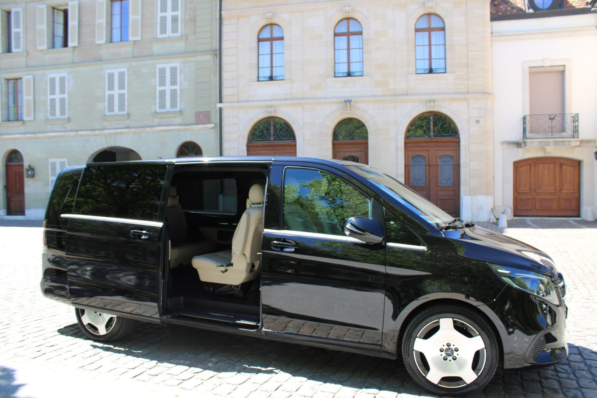 Une camionnette noire, porte latérale coulissante ouverte, est garée dans une rue pavée, devant des immeubles.