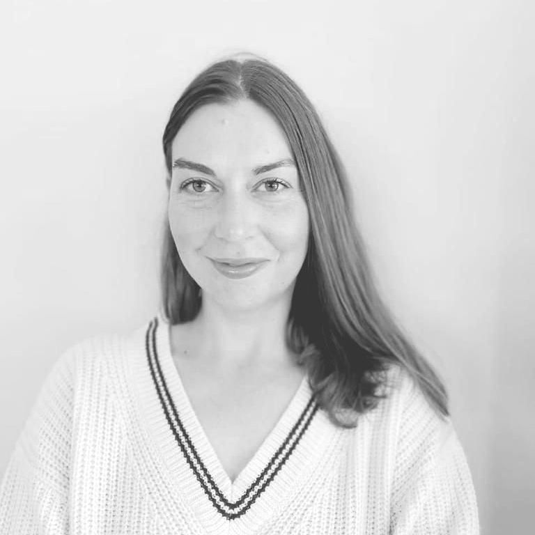 Willemien Froneman with long brown hair, wearing a white sweater with black trim, smiling, against a white wall.
