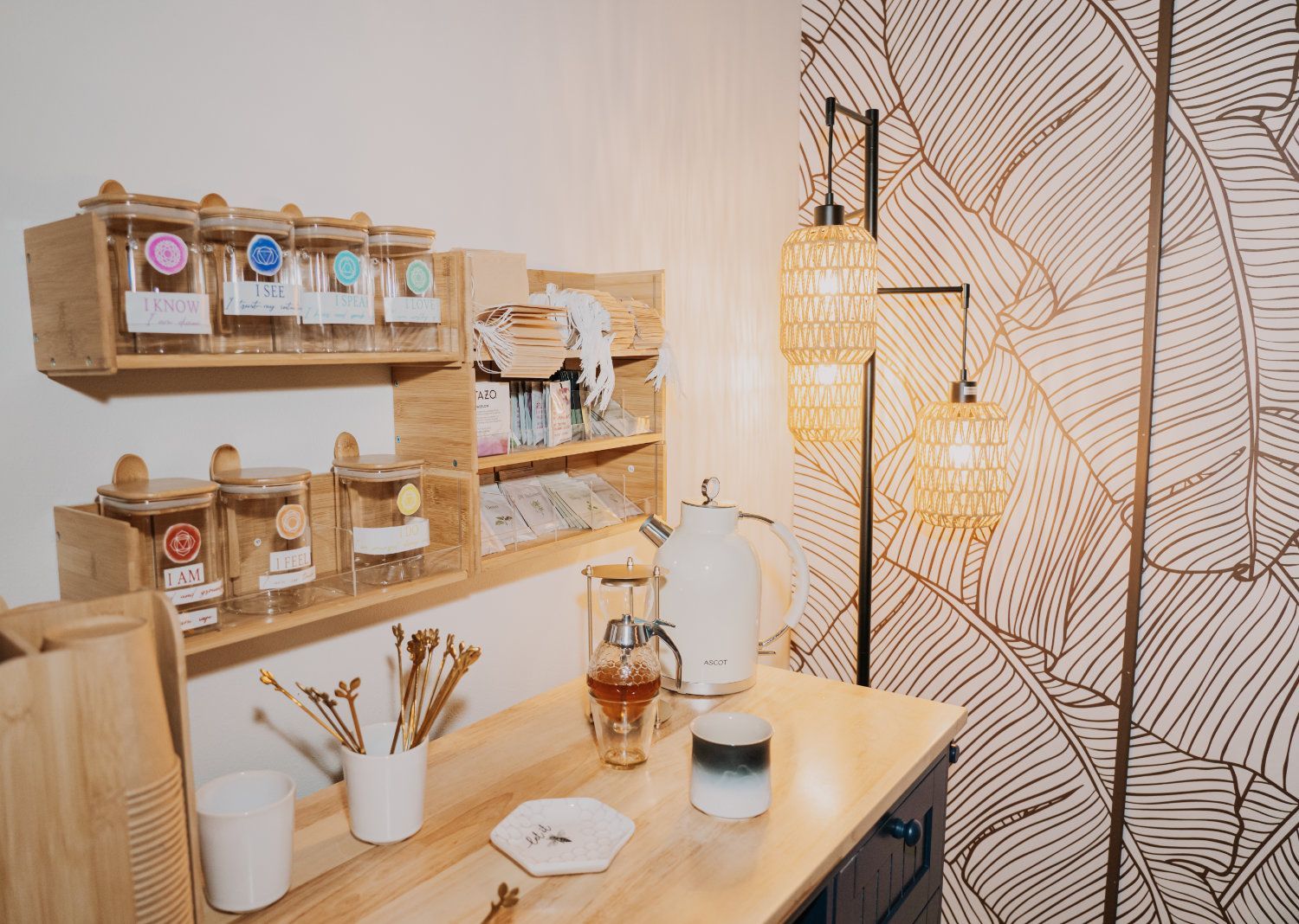 A cozy tea station with wooden shelves holding tea supplies, a lamp, and patterned wallpaper.