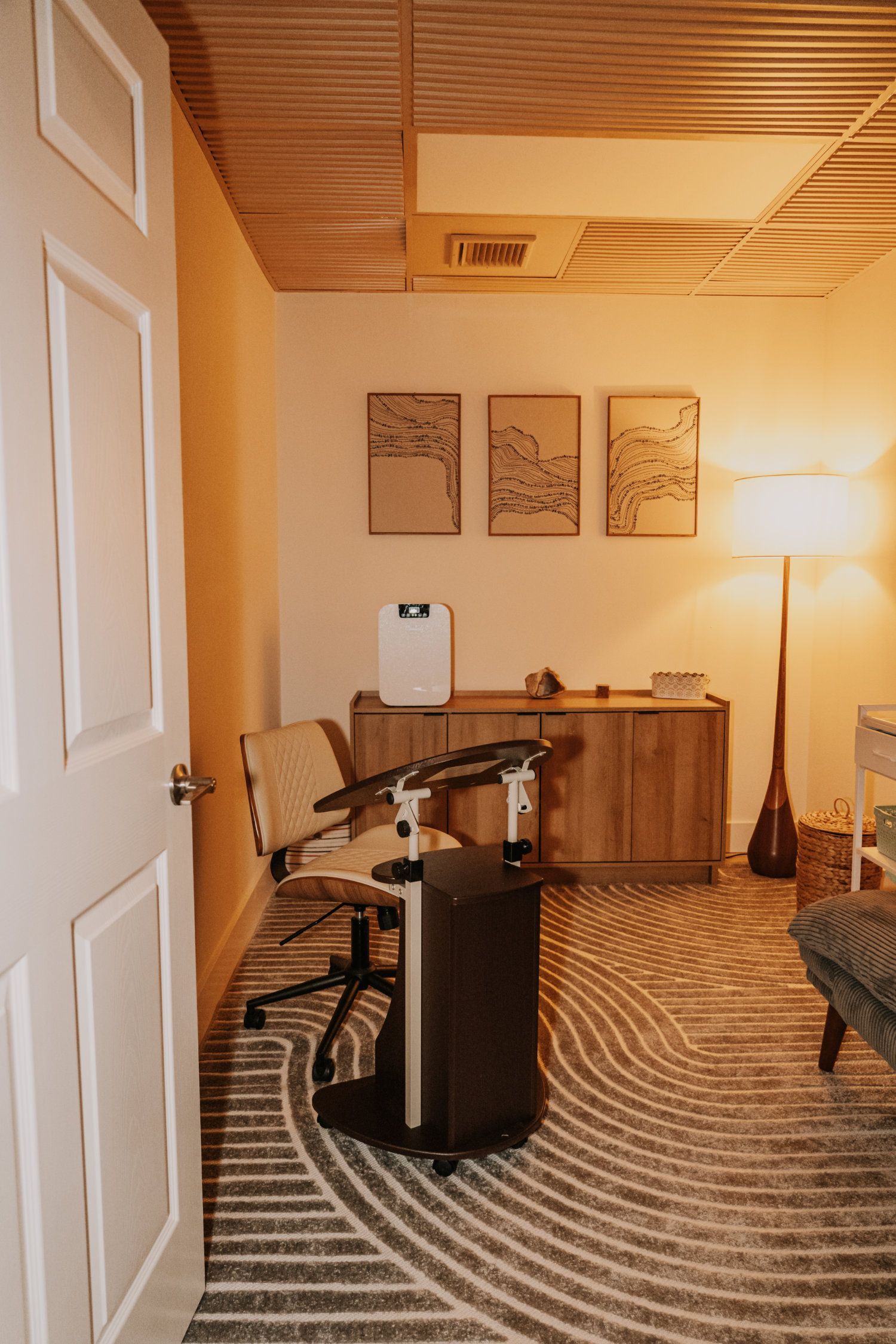 A therapy room with neutral tones. A desk chair, art, cabinet, and lamp on a patterned rug.