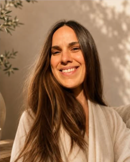 Smiling woman with long brown hair in a light sweater, standing indoors against a warm beige wall.