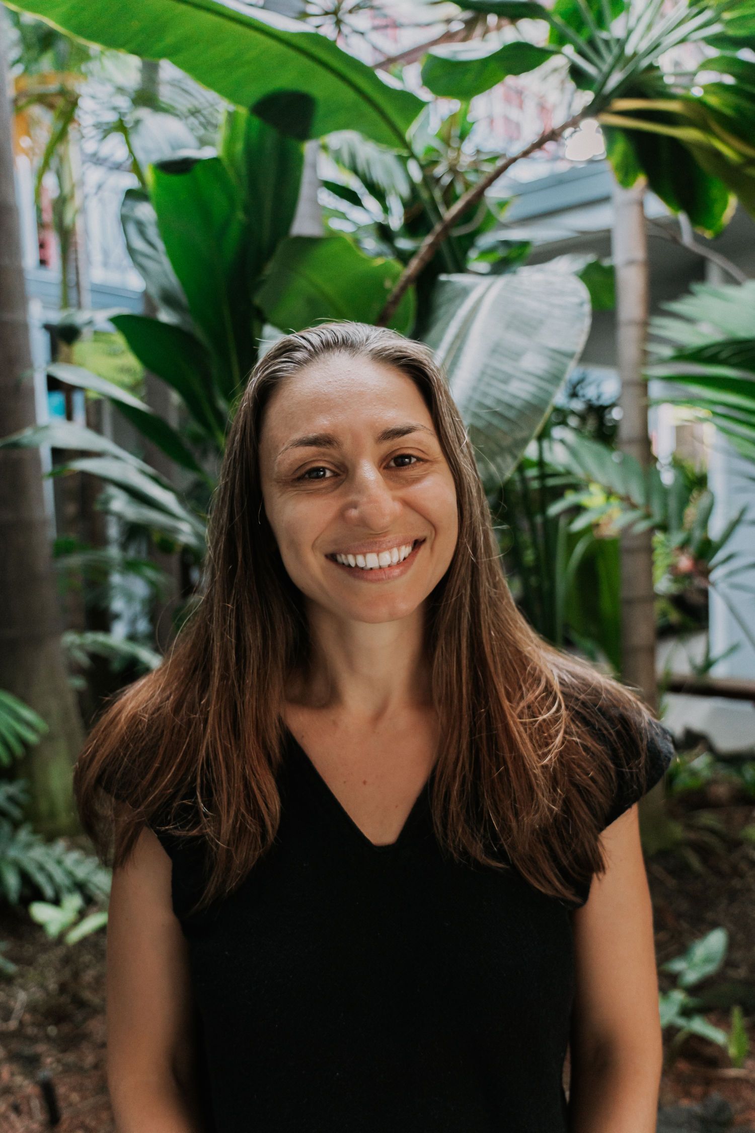 Woman with long brown hair smiles in front of lush green plants.
