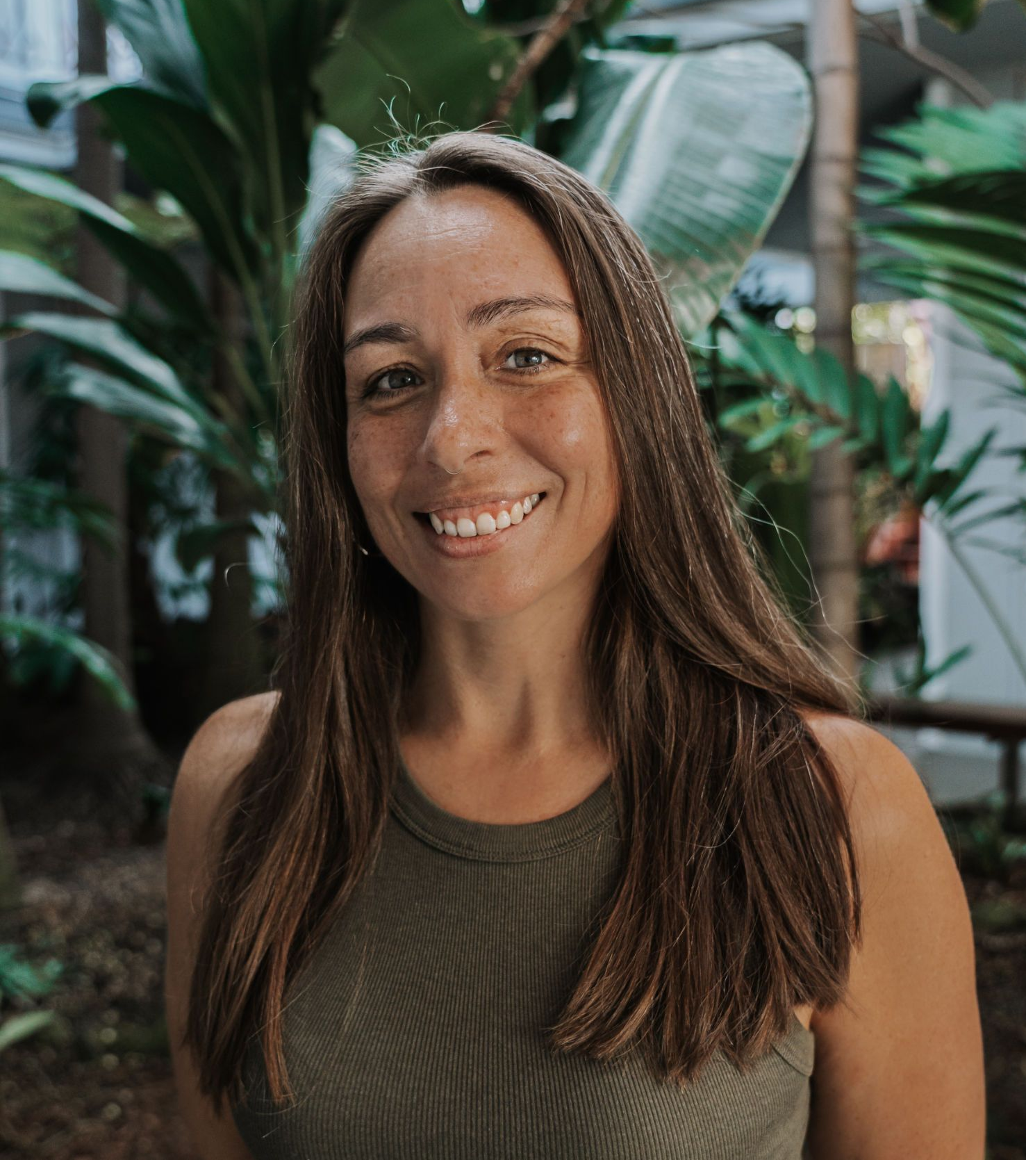 Woman with long brown hair smiles at the camera, wearing a green tank top, in a lush, green setting.