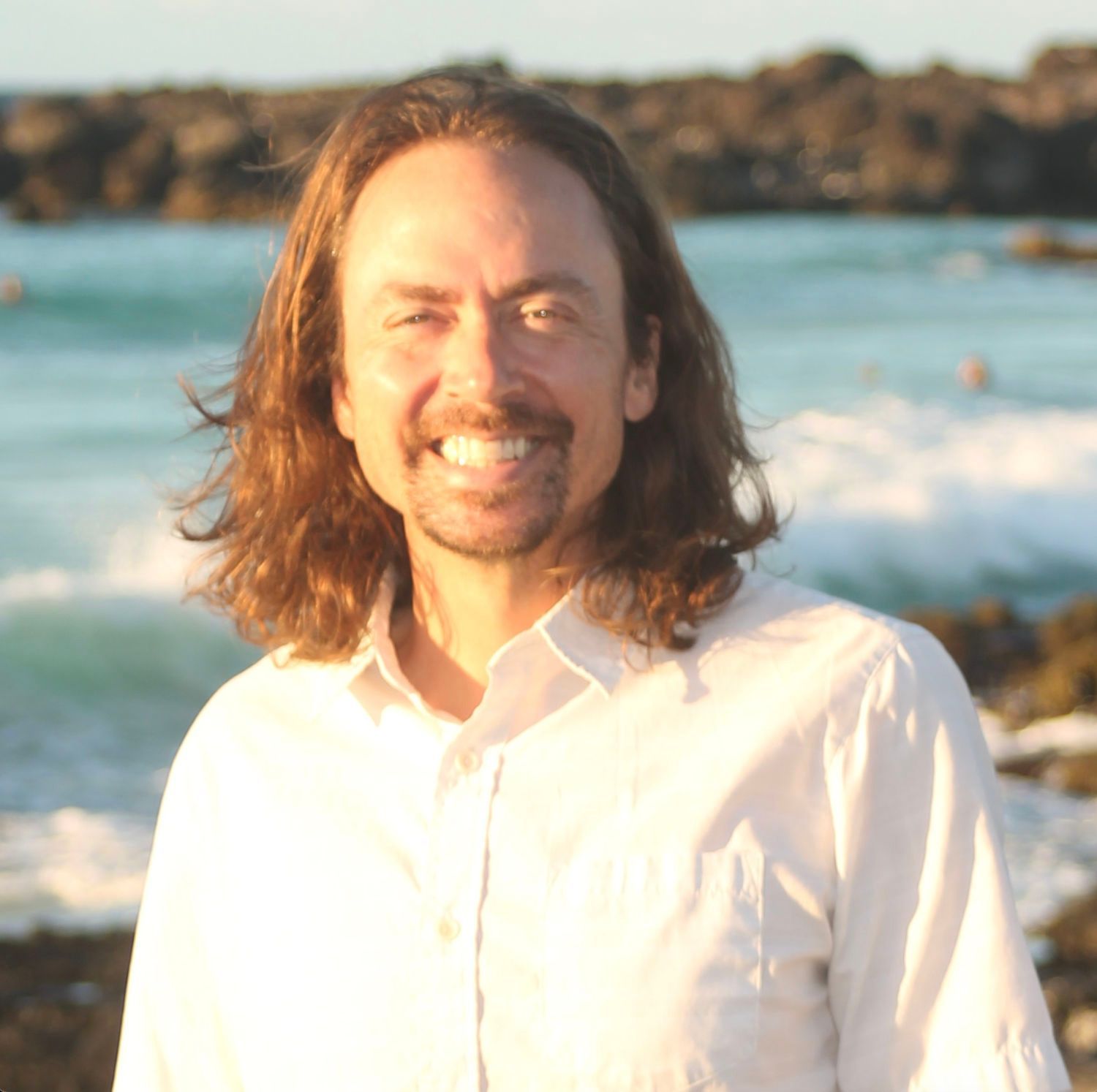 Smiling person in a white shirt standing by the ocean at sunset