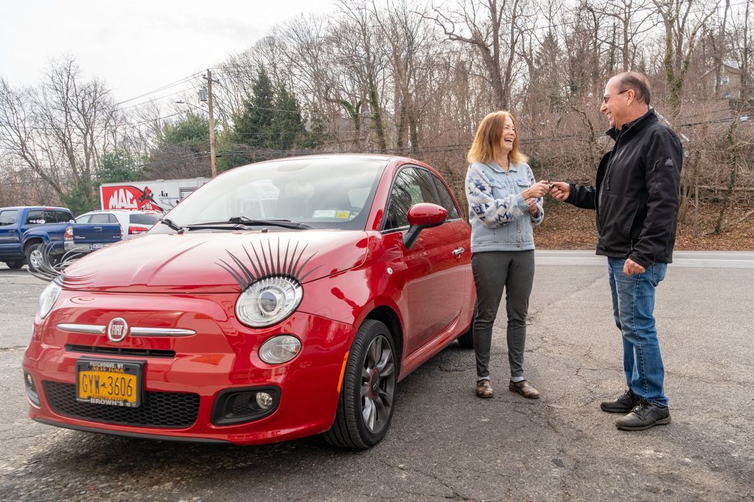 Man handing car keys to a woman near a red Fiat, parked outdoors.