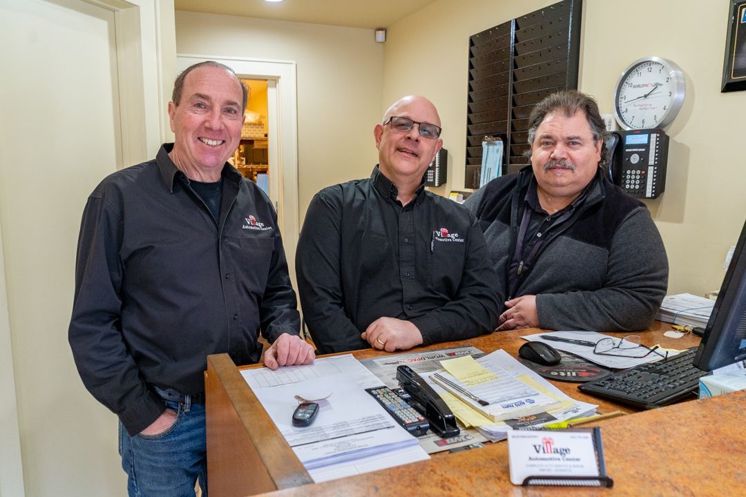 Three men stand behind a counter in an office. They smile, one wearing jeans, the others black shirts.