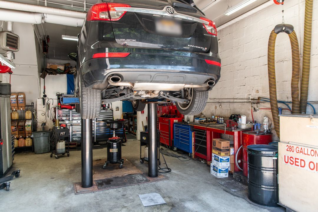 Car lifted on a garage hoist inside an auto repair shop, with tools and oil drums nearby.