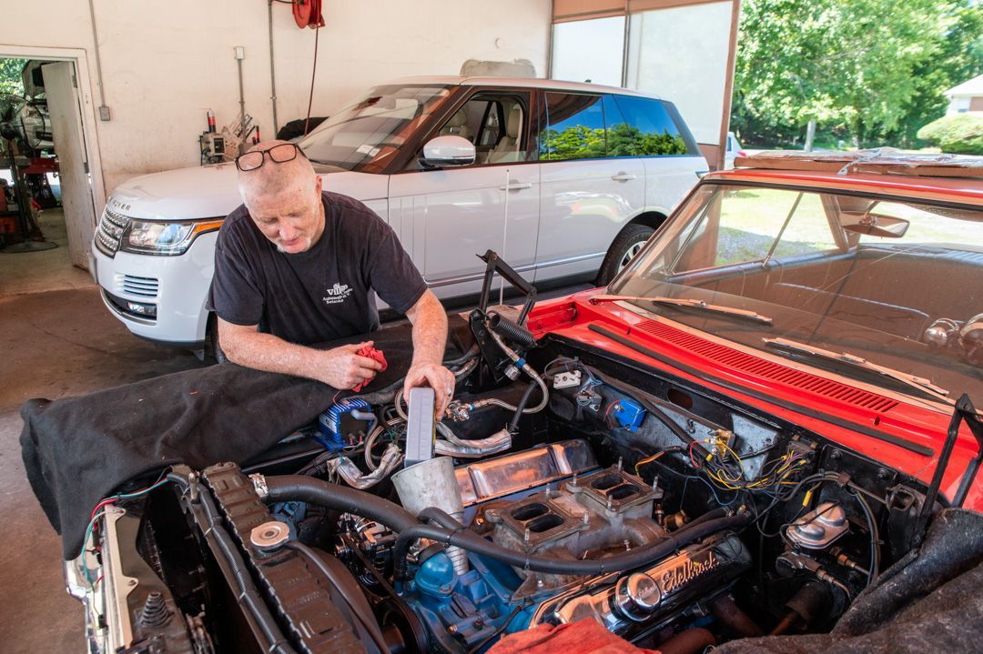 Man working on car engine in a garage, with a silver car parked behind him.