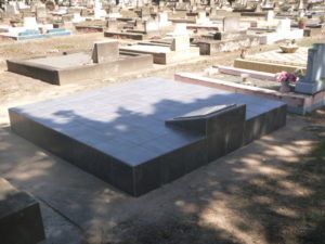 Large, flat, gray headstone in a cemetery with other headstones in the background.