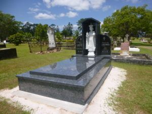Dark granite tombstone with statue of a figure, in a grassy cemetery setting under a partly cloudy sky.