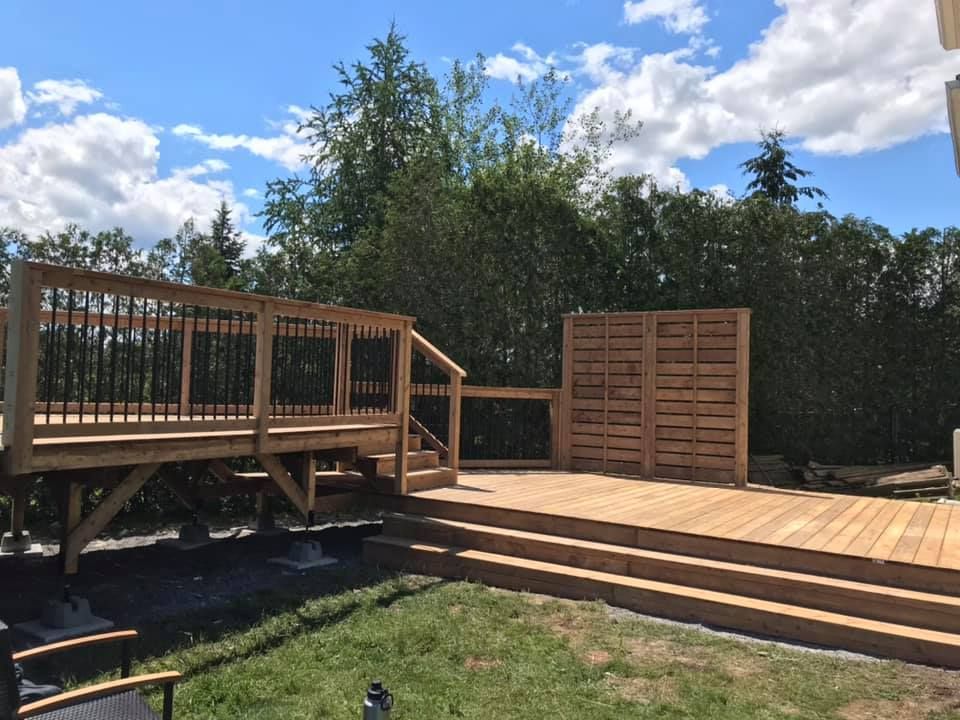 Une terrasse en bois à plusieurs niveaux dans le jardin, avec des balustrades en métal noir, un large escalier et un écran d'intimité, le tout sur fond de forêt.