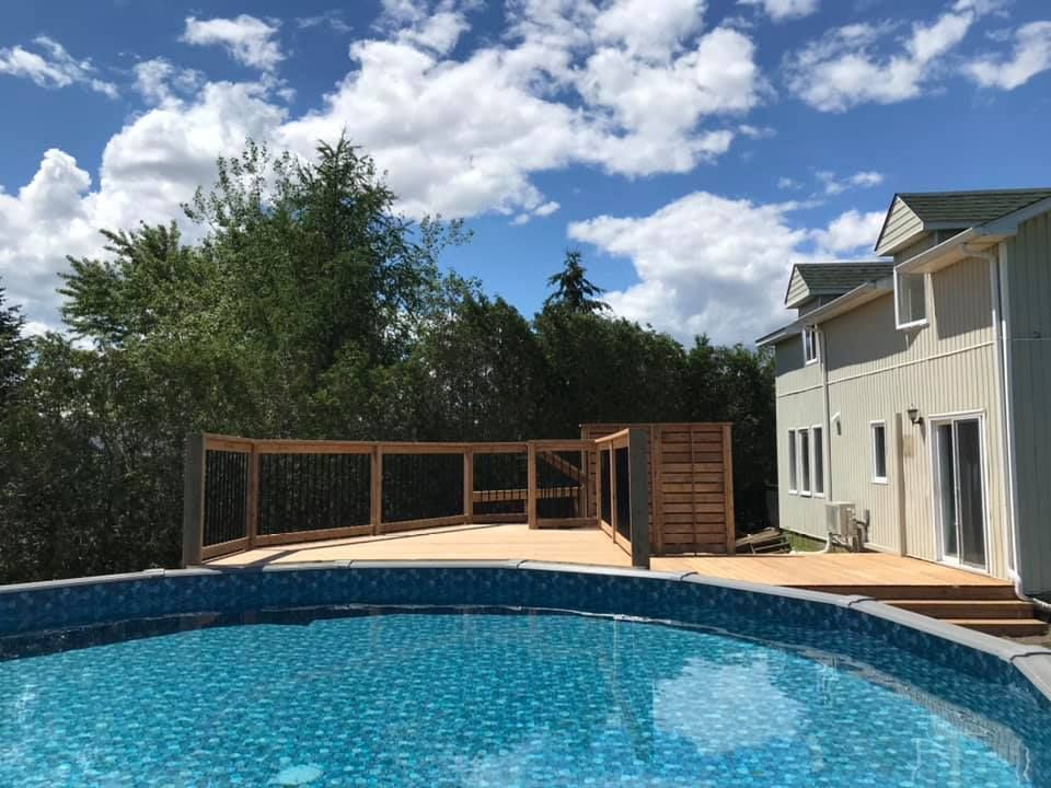 Une piscine hors-sol ronde se trouve devant une terrasse en bois et une maison beige à deux étages, sous un ciel bleu nuageux.