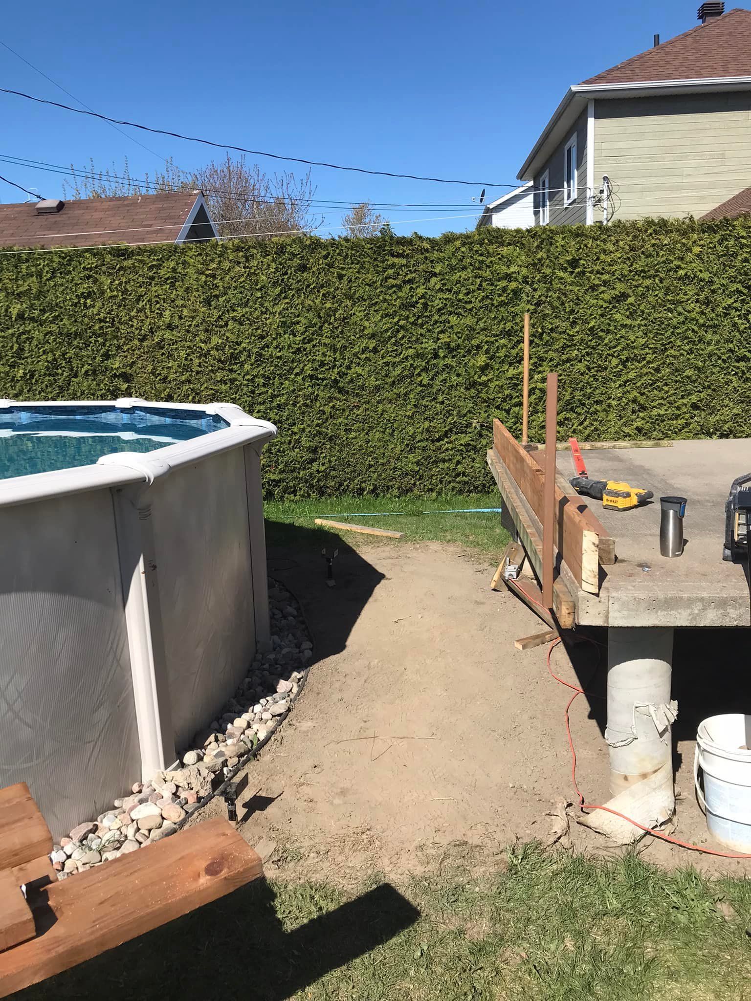 Une piscine hors sol jouxte une terrasse inachevée et un chemin de terre dans un jardin résidentiel bordé d'une haute haie.
