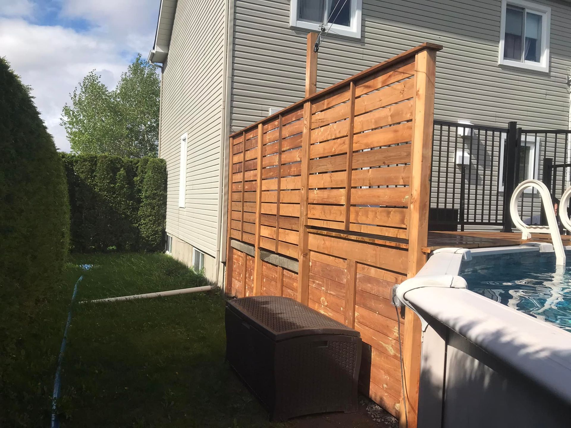 Une scène de jardin montrant une haute clôture en bois pour préserver l'intimité, à côté d'une piscine hors sol et d'un coffre de rangement marron.