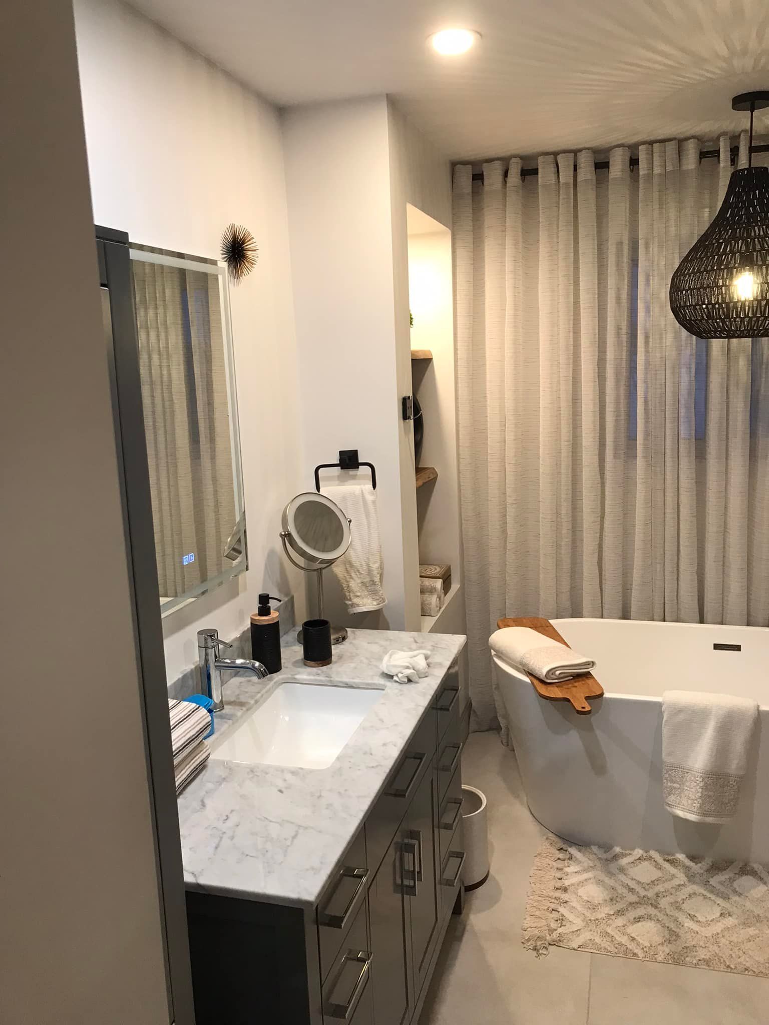 Une salle de bains avec un meuble-lavabo en marbre gris, un miroir éclairé, une baignoire blanche et des rideaux blancs transparents.