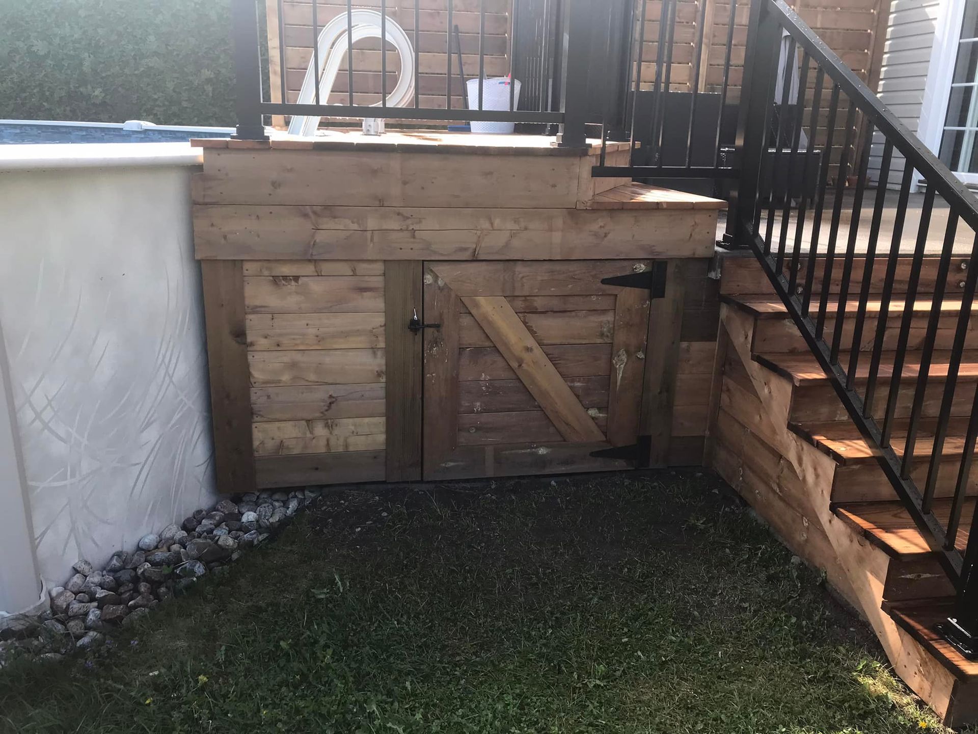 Une terrasse basse en bois avec une porte de rangement intégrée en dessous, située à côté d'un escalier et d'un mur de piscine de couleur claire.