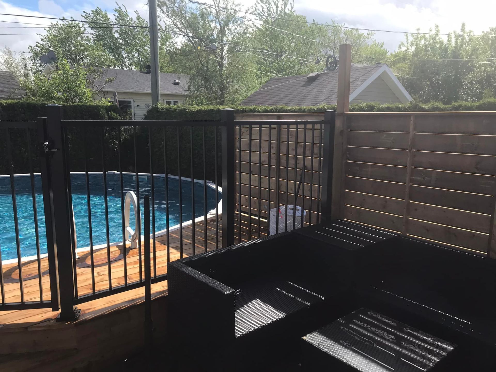 Un canapé d'extérieur en osier noir est installé sur une terrasse jouxtant une piscine entourée d'une clôture en métal noir et d'un muret en bois préservant l'intimité.