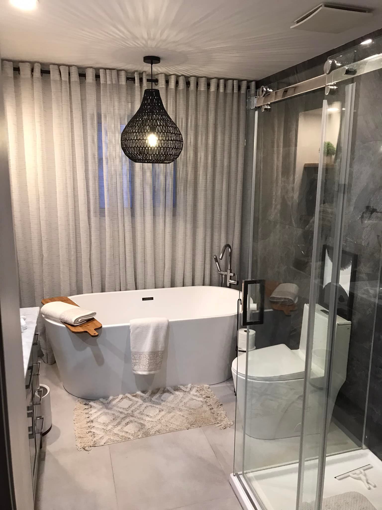 Salle de bains moderne avec baignoire îlot, douche à l'italienne en verre, suspension décorative et rideaux allant du sol au plafond.