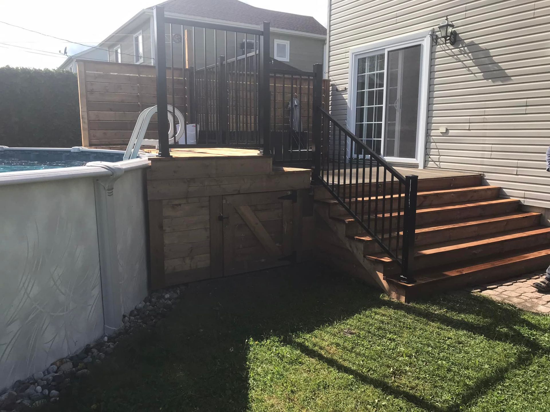 Une terrasse en bois avec des balustrades noires relie une porte coulissante de la maison à une piscine hors sol située dans un jardin gazonné.