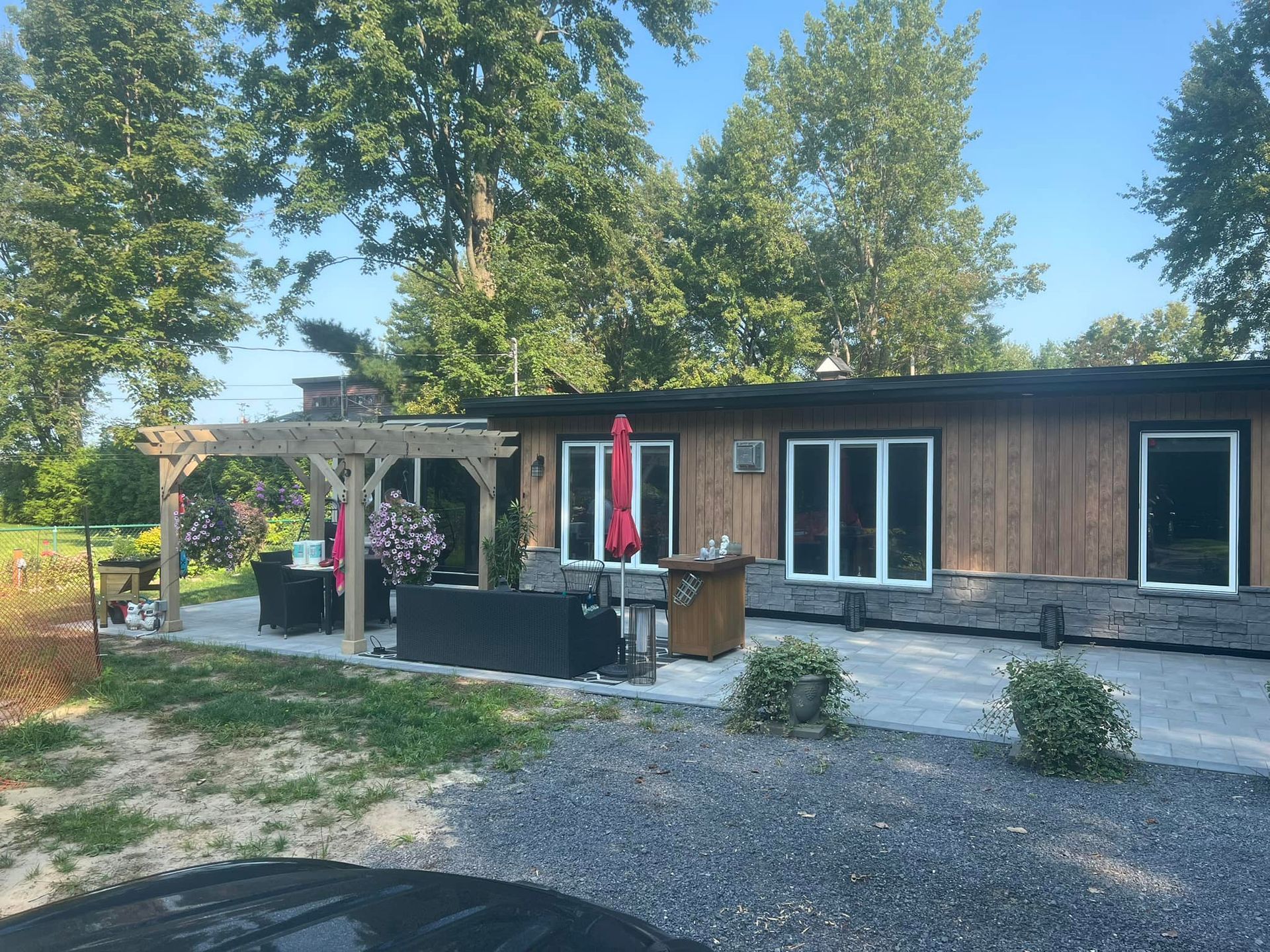 Une maison en bois avec des fondations en pierre et une terrasse en pierre aménagée avec du mobilier de jardin, une pergola et une zone gravillonnée.