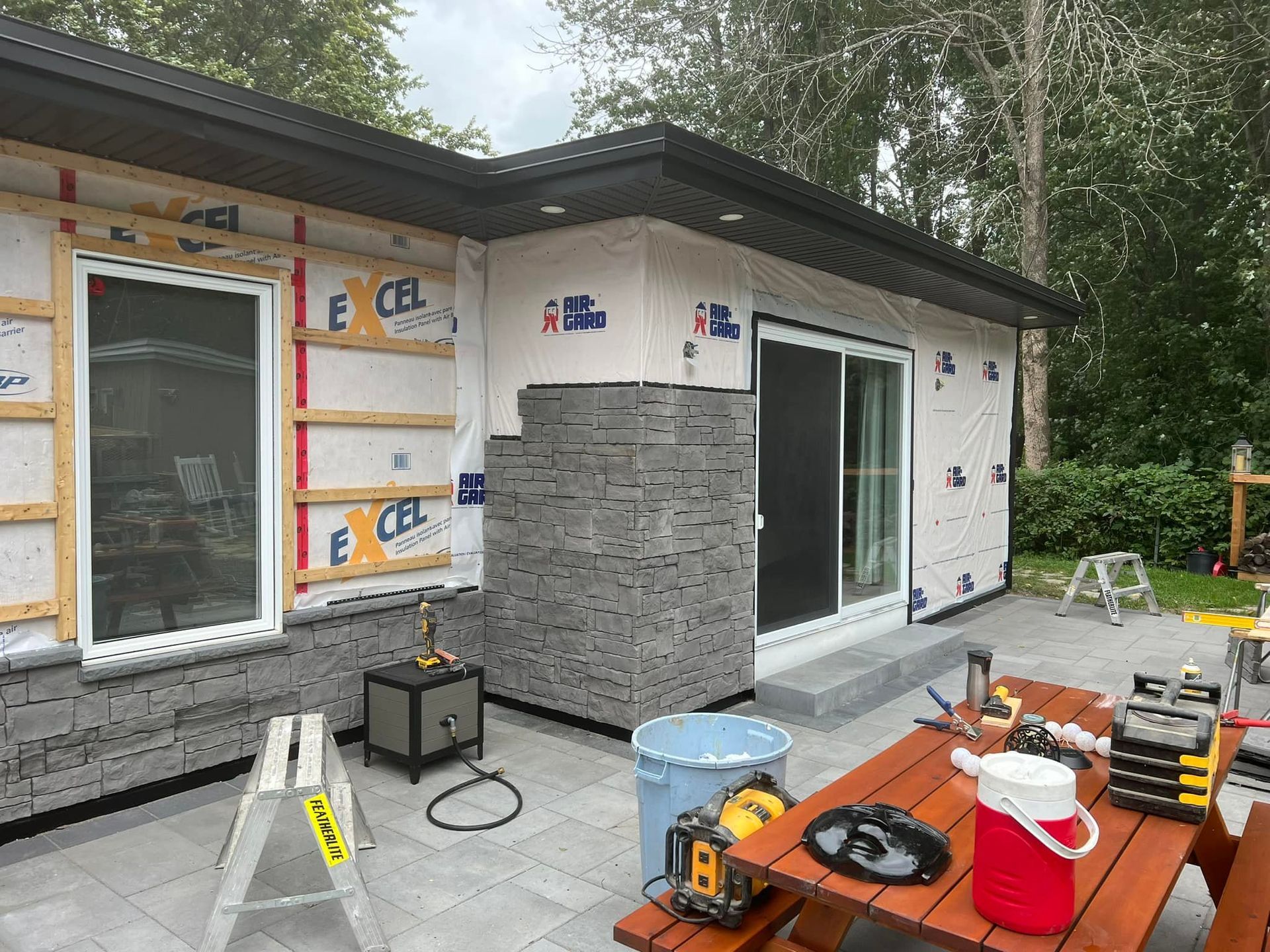 Rénovation d'un mur extérieur avec parement en pierre partiellement installé sur une maison, laissant apparaître l'isolation et les outils sur une terrasse.