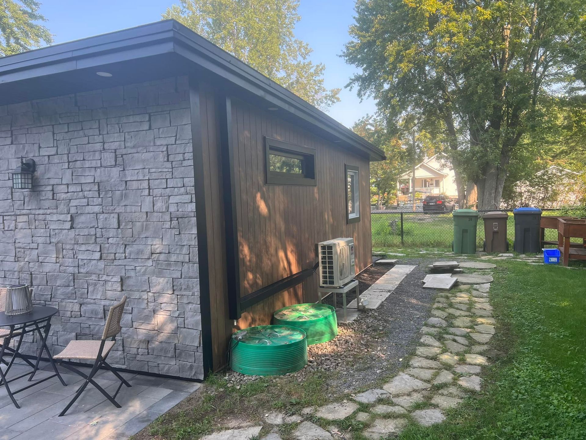 Une structure de jardin à l'aspect pierreux avec un mur latéral en bois foncé, une unité de climatisation, un chemin en pierre et des bacs verts dans une cour gazonnée.