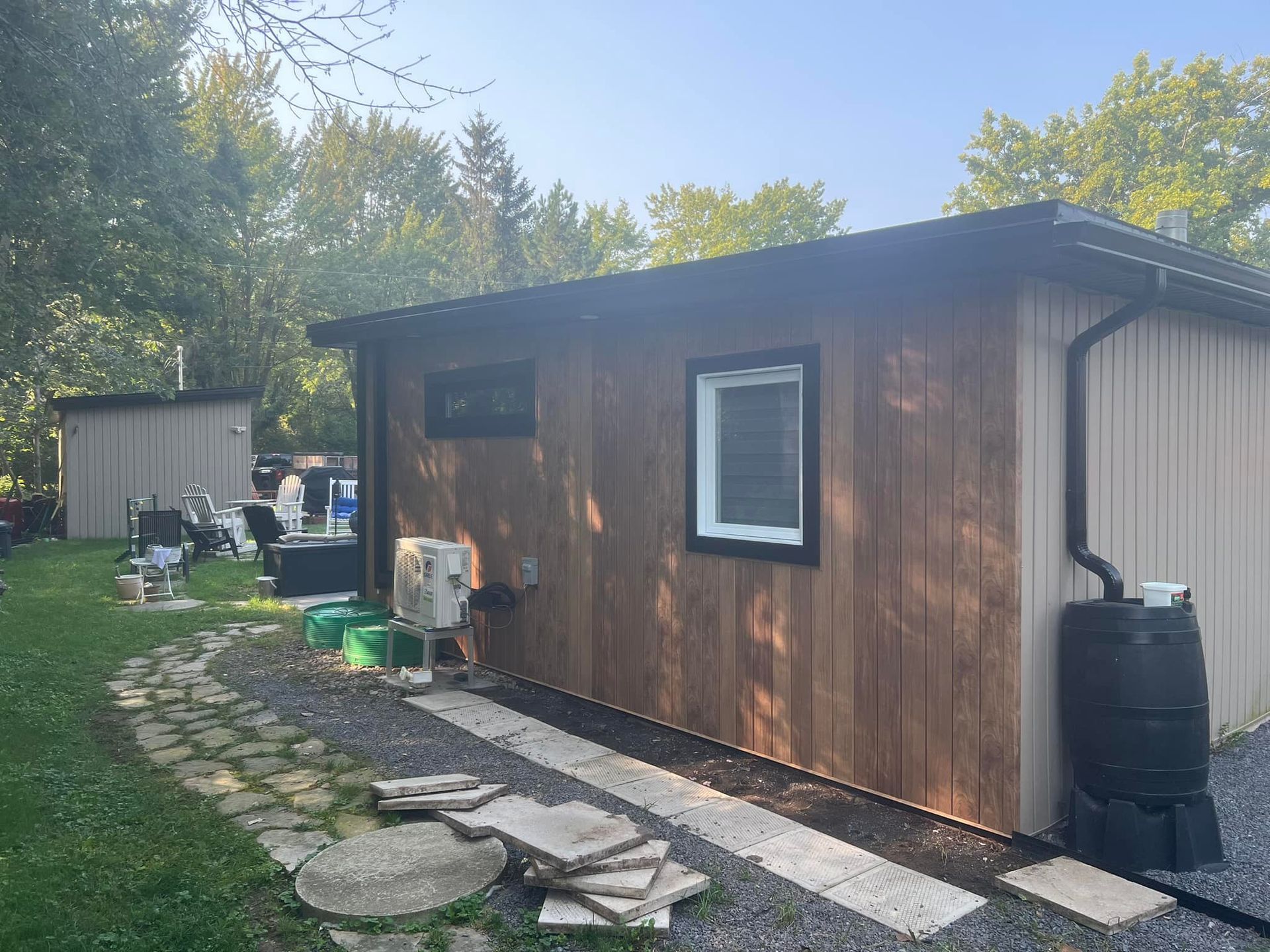Une cabane en bois avec des boiseries et un toit noirs, dotée d'une fenêtre et d'un récupérateur d'eau de pluie, située sur un chemin de gravier dans une cour herbeuse.