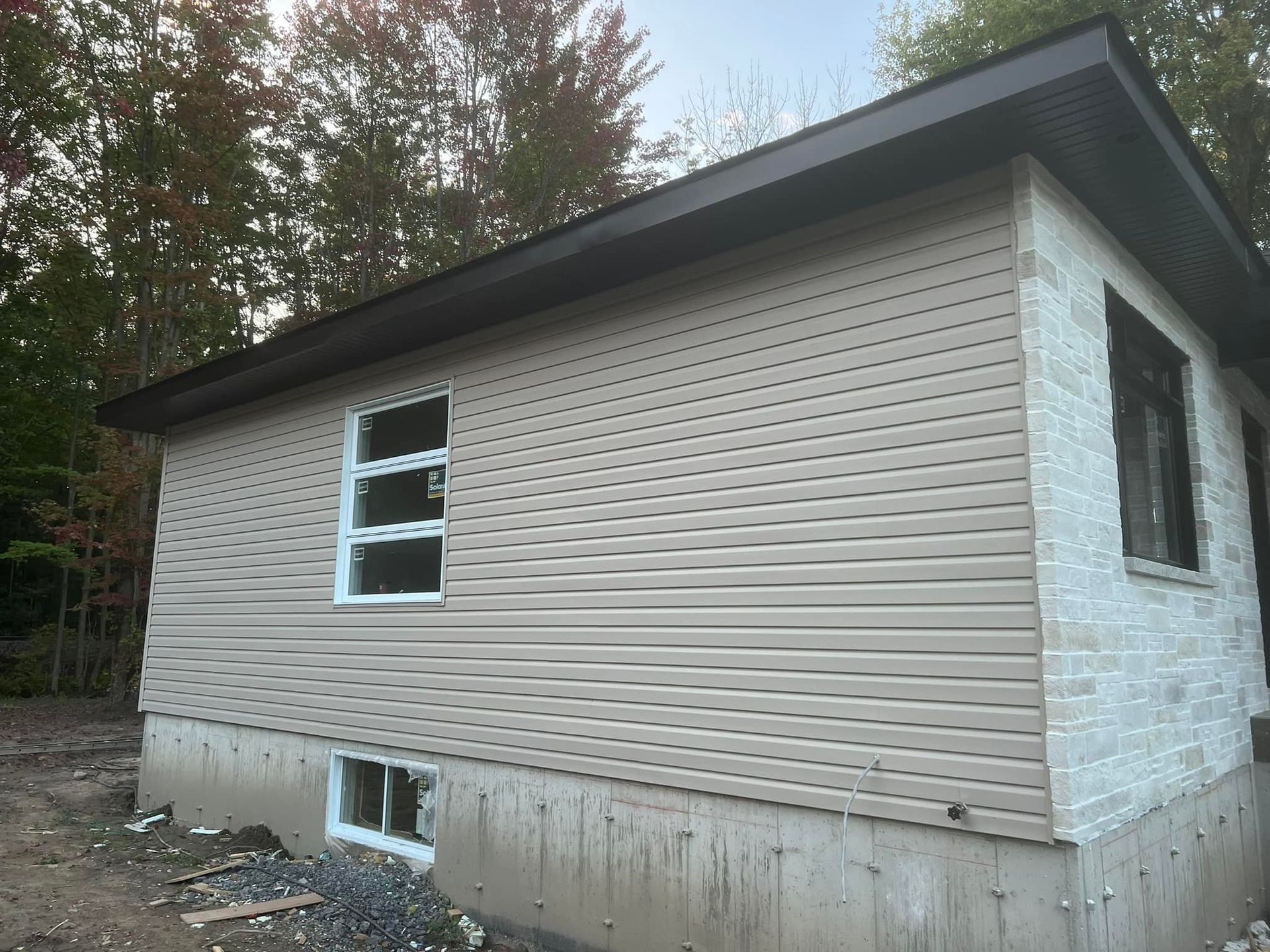 Une maison en construction avec un bardage horizontal beige, un angle en pierre blanche et des fondations en béton avec deux fenêtres.