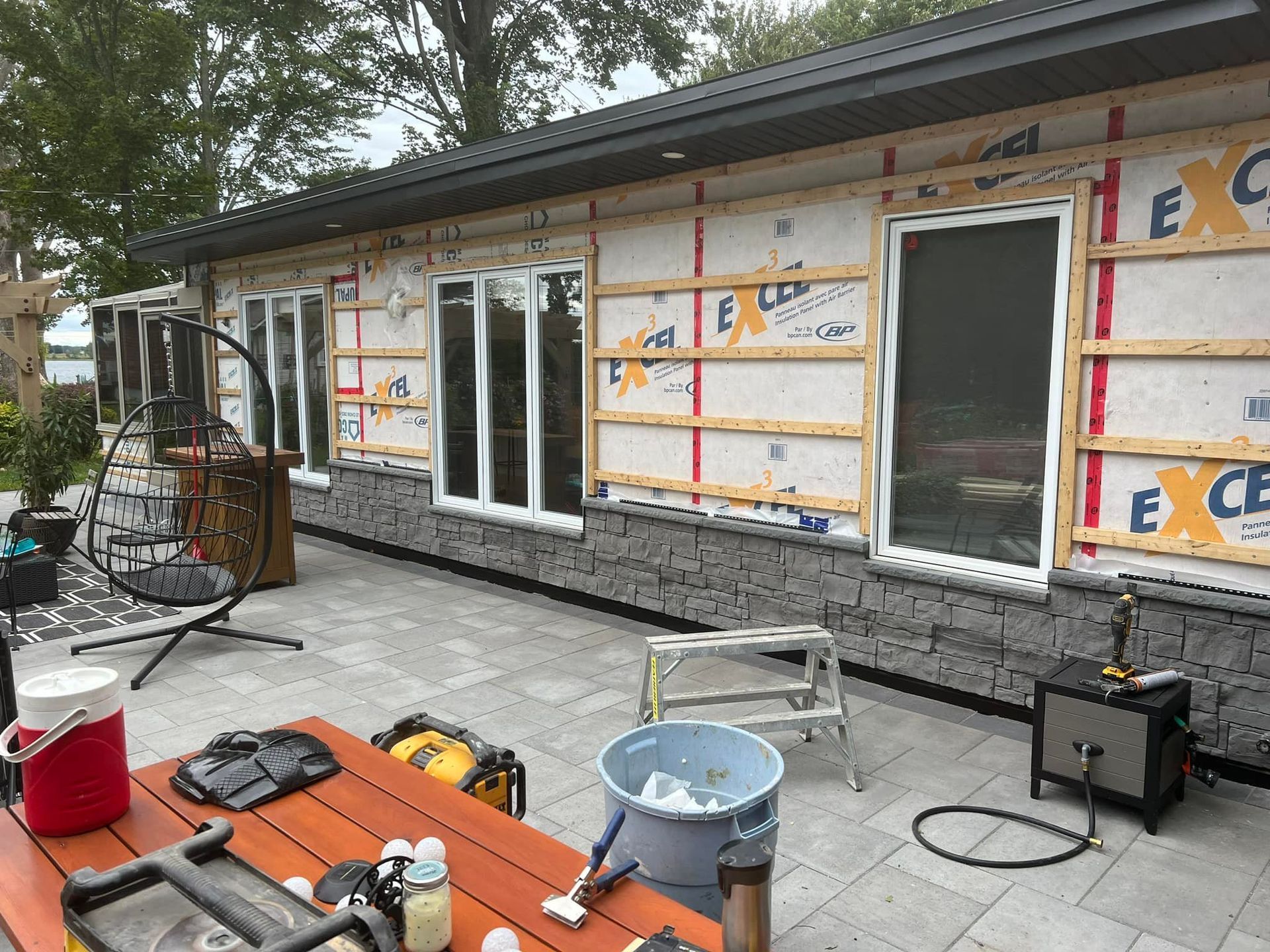 L'extérieur d'une maison en construction, avec une ossature en bois horizontale installée sur un pare-pluie blanc et un revêtement en pierre grise.