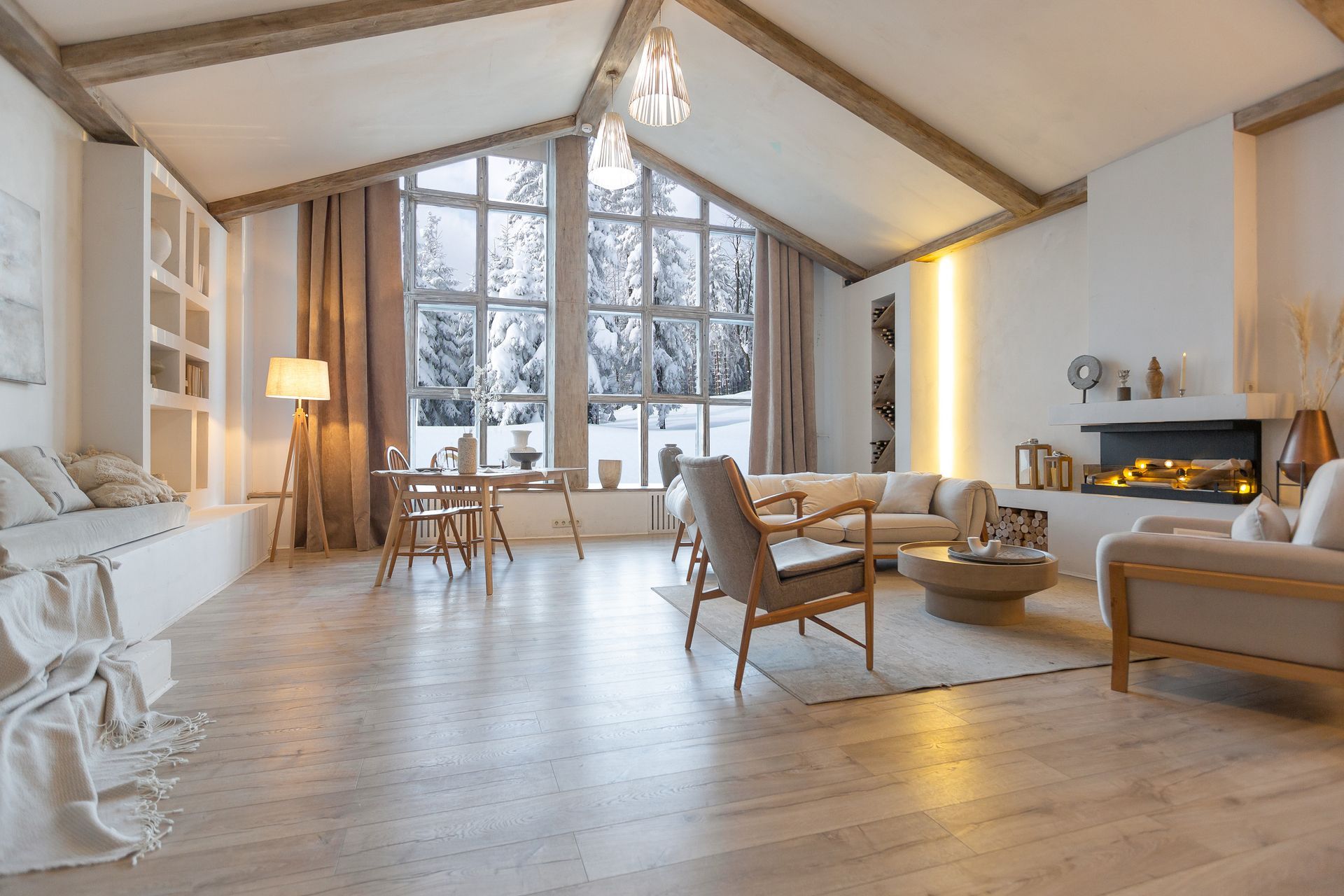 Un salon chaleureux et lumineux, avec des poutres apparentes, une cheminée et de grandes fenêtres donnant sur un paysage forestier enneigé.