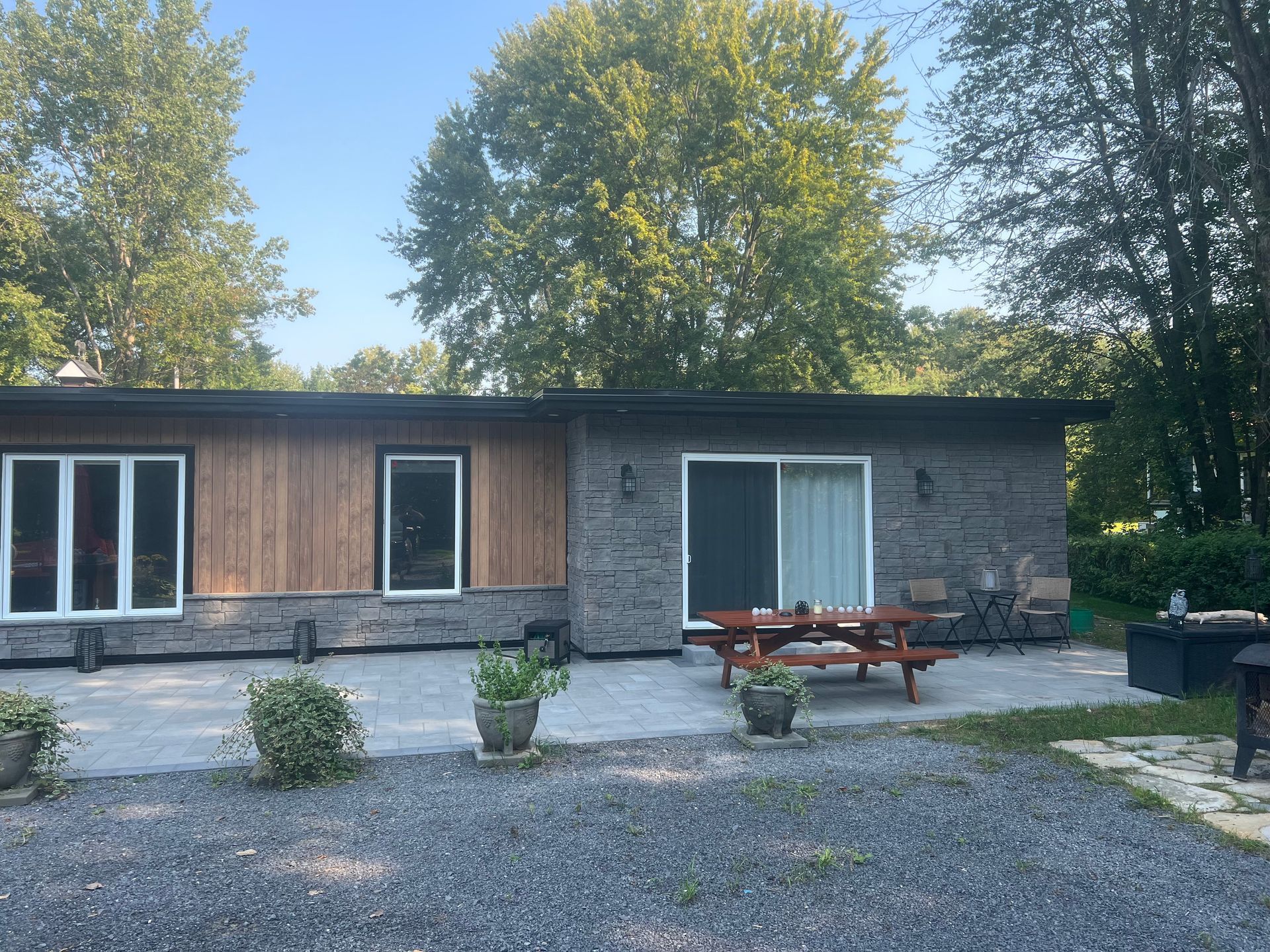 Une maison moderne de plain-pied avec un revêtement en bois et en pierre, une terrasse avec une table de pique-nique et une cour gravillonnée sous un ciel bleu.