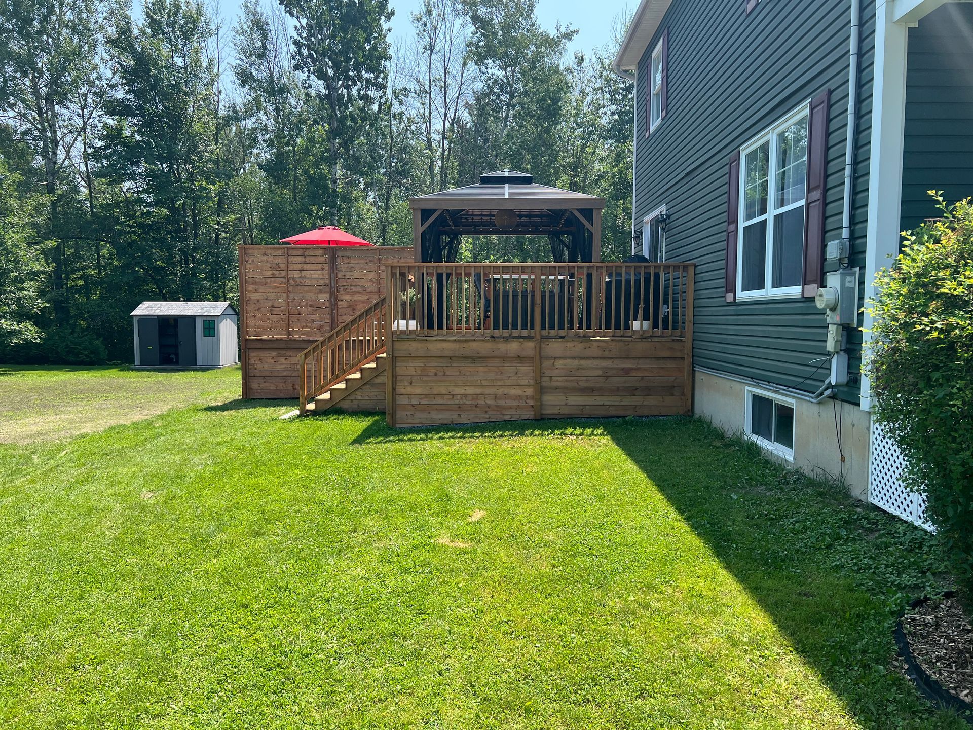 Une terrasse en bois à l'arrière d'une maison aux murs sombres, avec un kiosque et un parasol rouge, jouxte un petit abri de jardin et une pelouse.